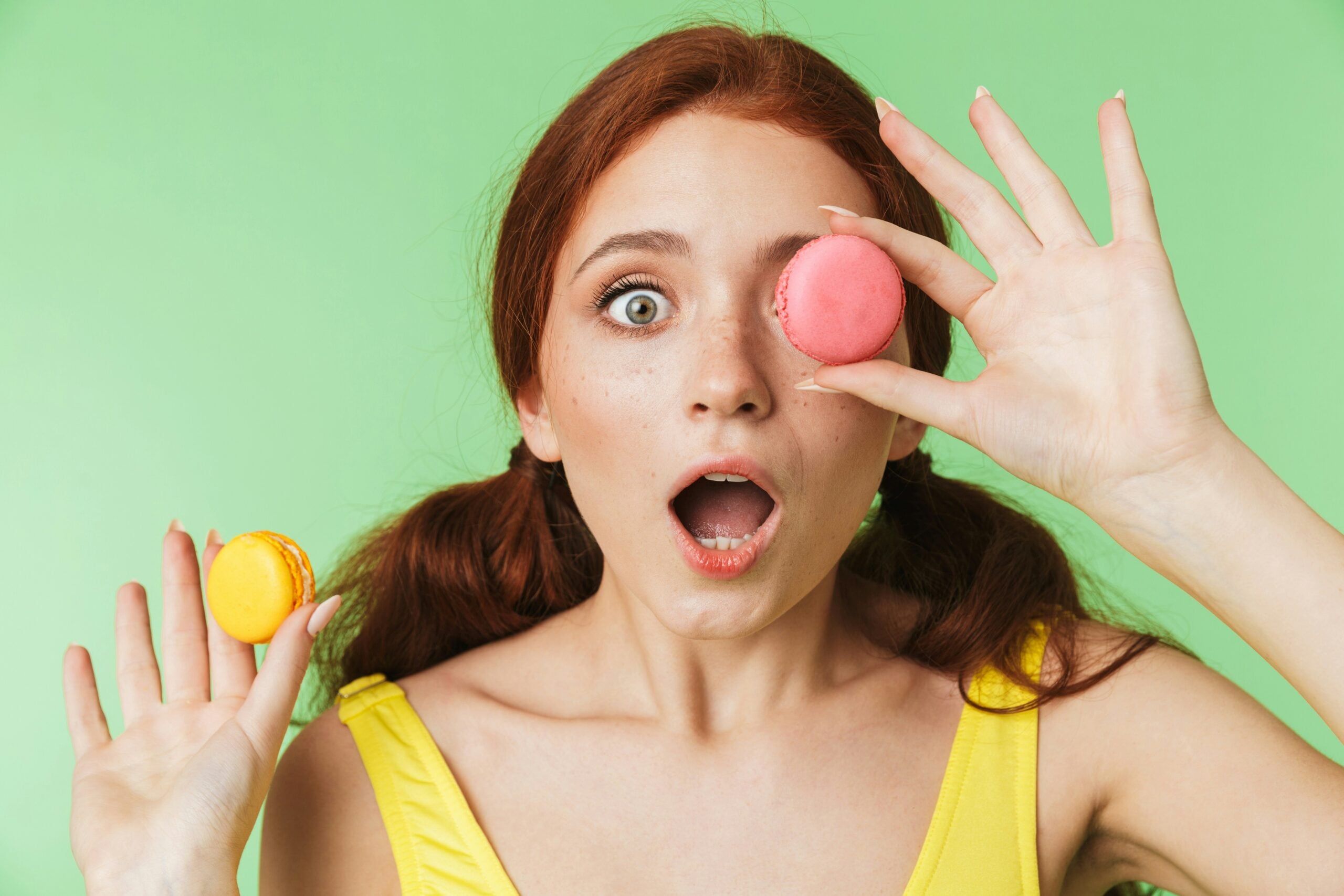 Surprised young woman with red hair holds a yellow macaron in one hand and covers one eye with a pink macaron, standing in front of a green background.