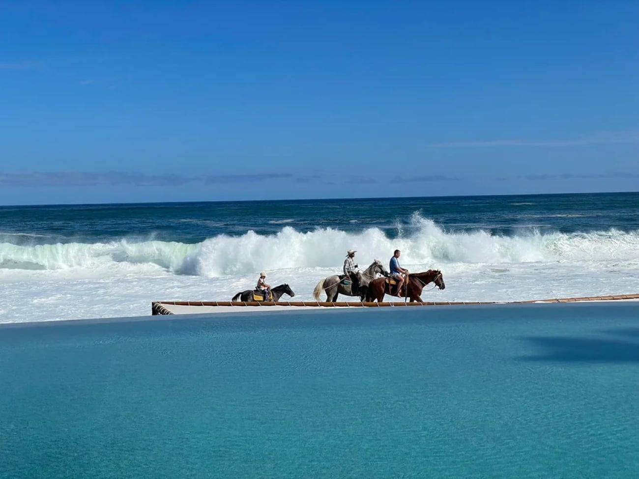 Three people riding horses along a sandy shore with crashing waves behind them, and a calm blue pool in the foreground under a clear sky evoke the serene beauty found in tropical locations.