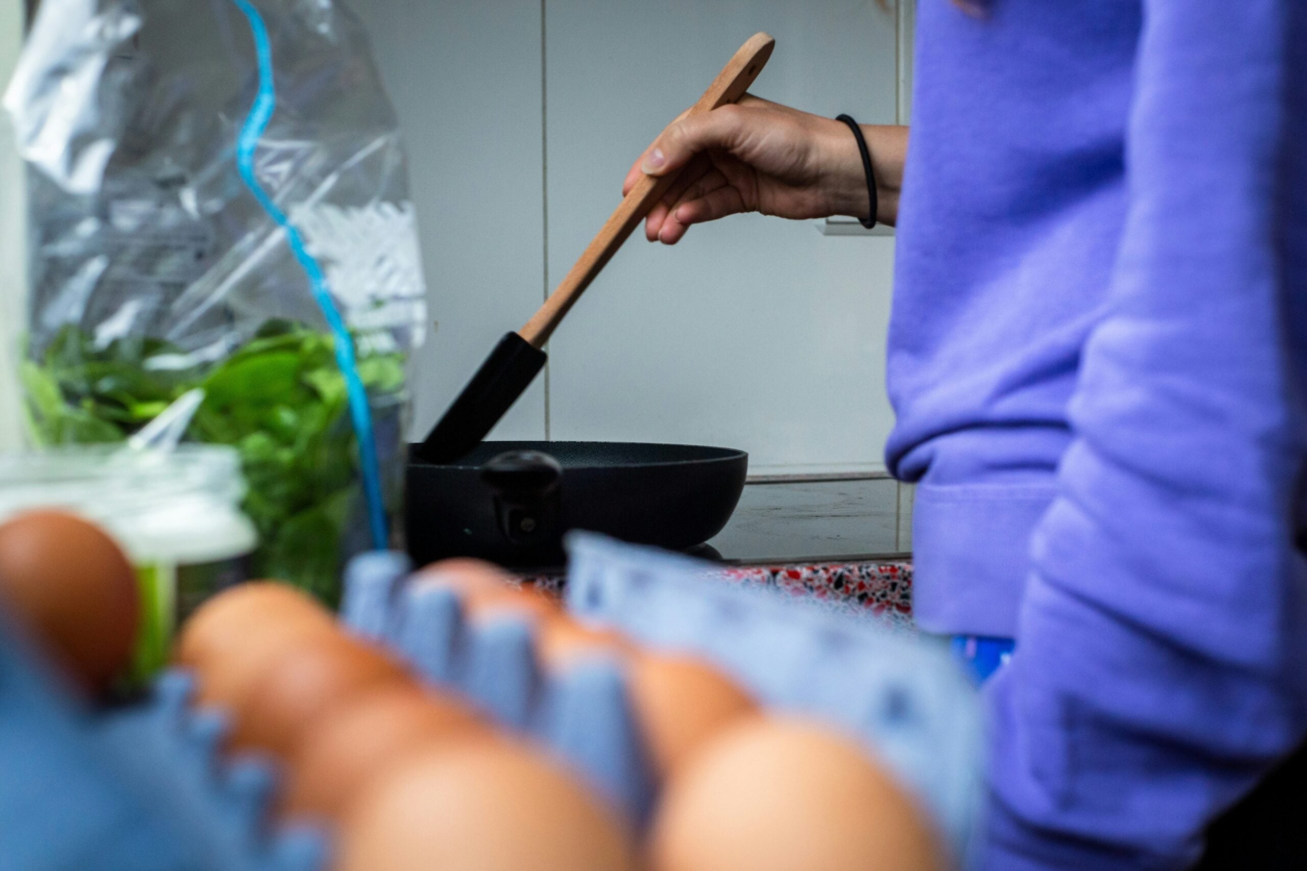 A person in a purple sweatshirt stirs a frying pan with a spatula. Eggs in a carton and green vegetables are on the counter in the foreground.