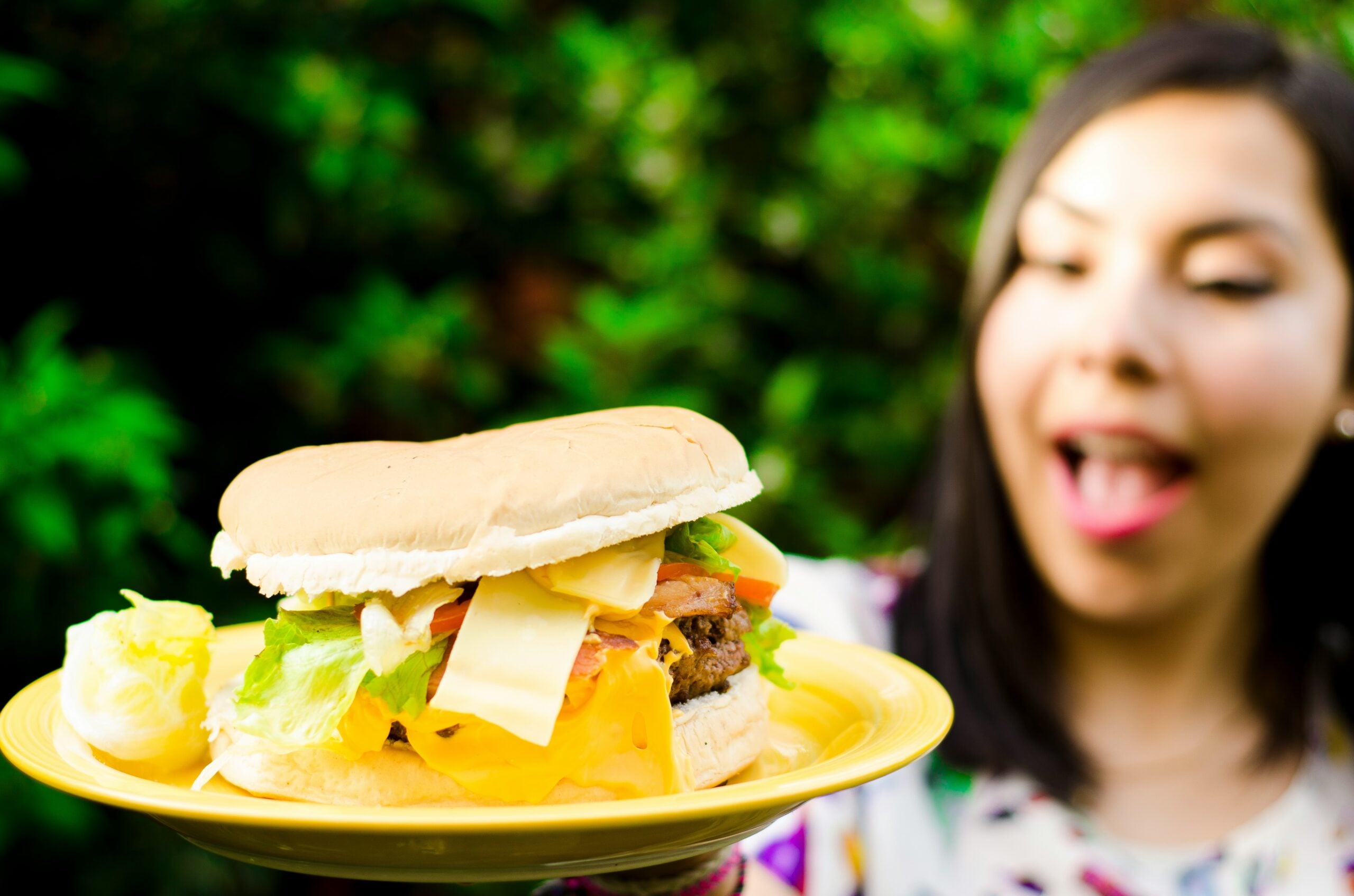 A woman with dark hair excitedly looks at a large cheeseburger with lettuce, tomato, and cheese on a yellow plate, with greenery blurred in the background.