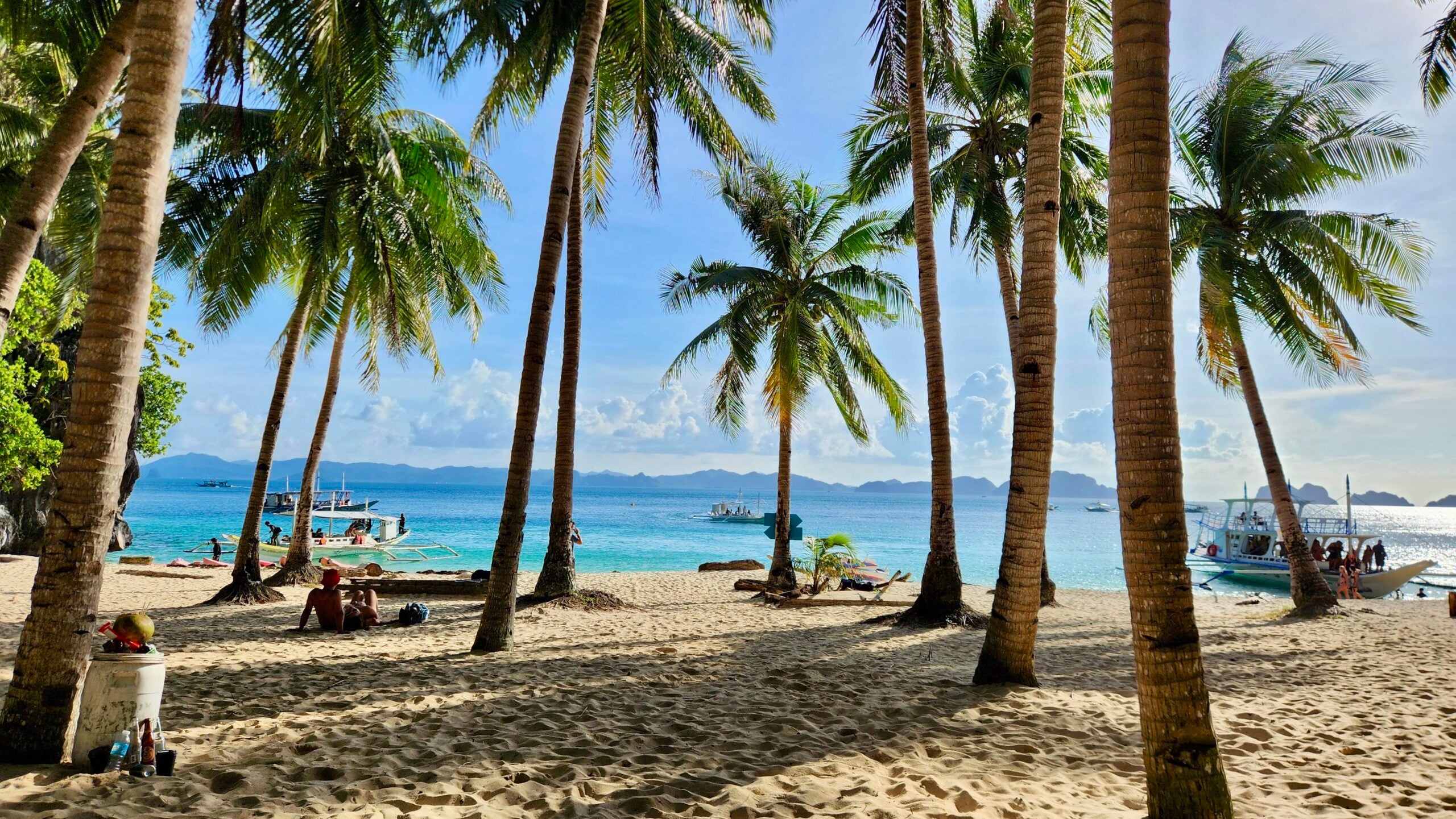 Tropical beach with palm trees, people lounging on the sand, boats on turquoise water, and distant islands under a bright blue sky—one of the region’s most incredible beaches.