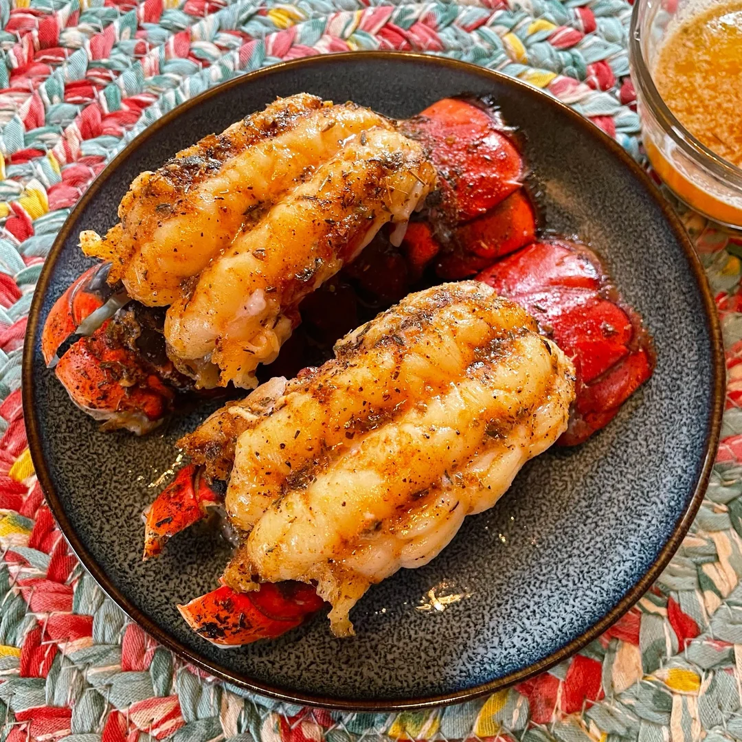 Two seasoned and broiled lobster tails served on a dark plate, sitting on a colorful woven placemat. A small glass of melted butter is visible in the top right corner.