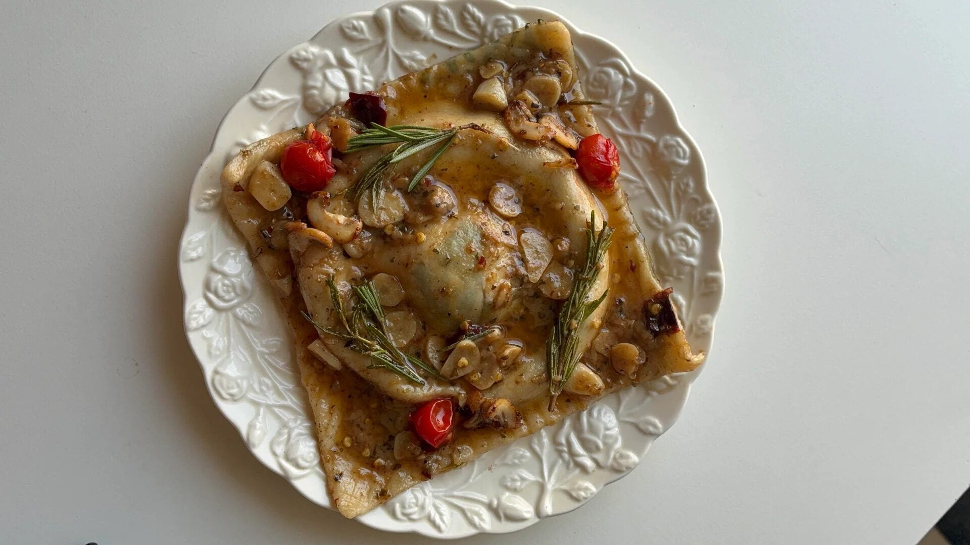 A large square raviolo topped with mushrooms, cherry tomatoes, herbs, and sauce sits on a decorative white plate against a plain background.