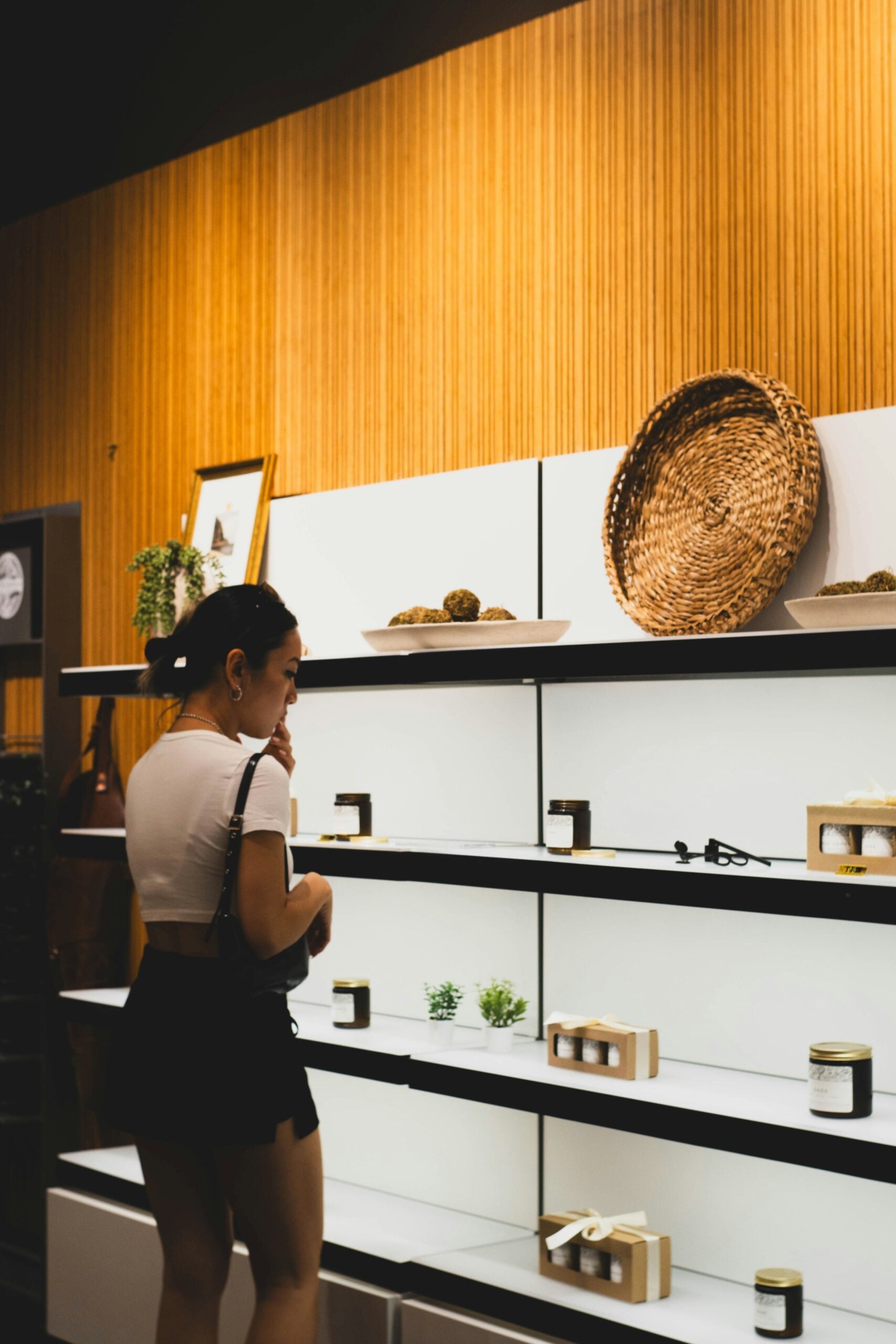 A woman stands in front of a modern shelf displaying jars, small plants, and a large woven basket against a wooden wall.