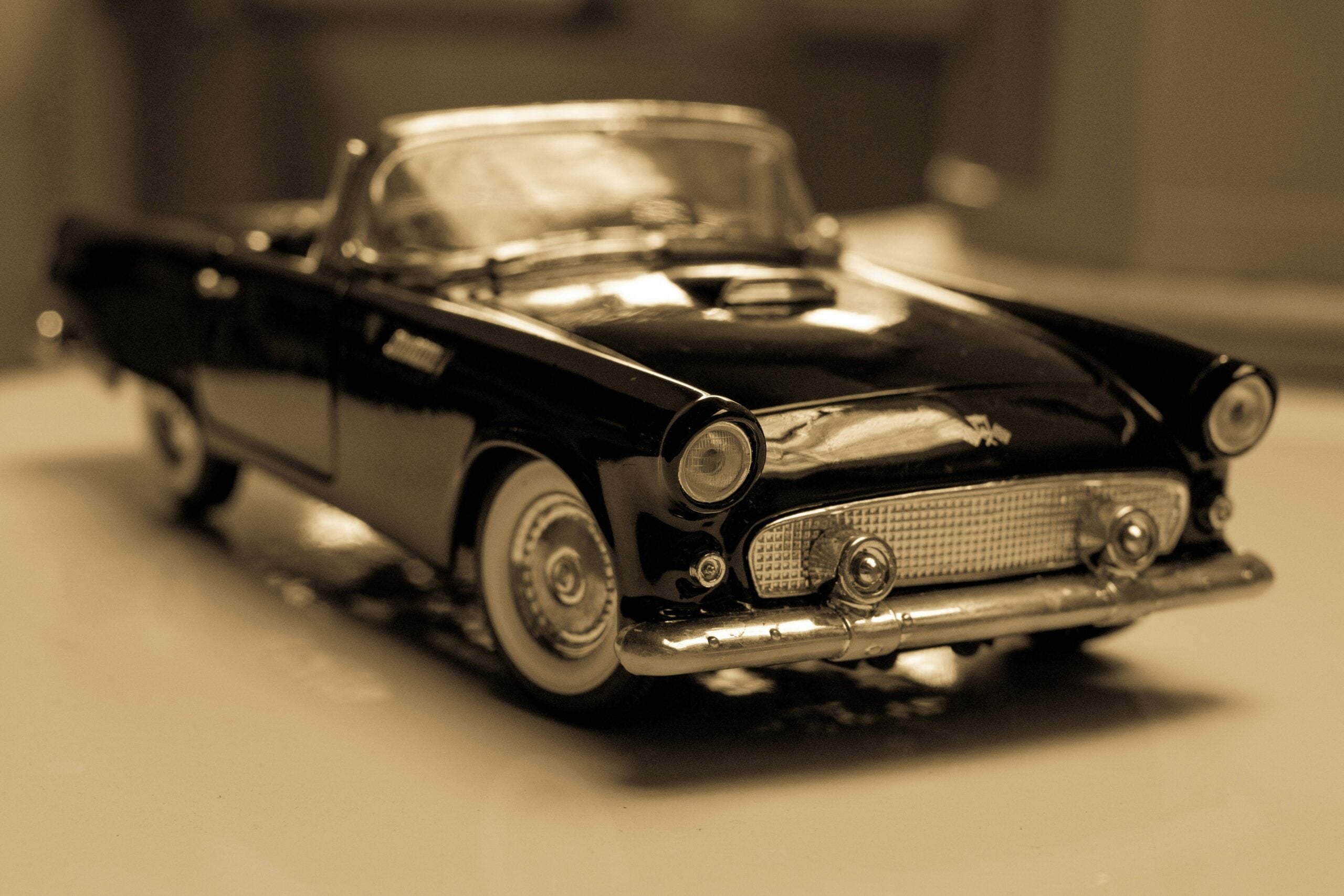 Sepia-toned close-up of a vintage convertible toy car, showcasing details like the chrome grille, headlights, and front bumper on a smooth surface.