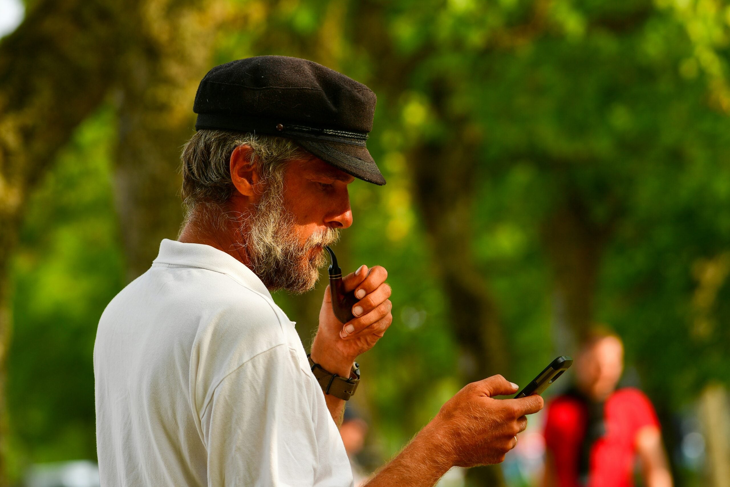 A bearded man in a white shirt and black cap holds a phone in one hand and touches his beard with the other, standing outdoors with trees blurred in the background, perhaps contemplating his retirement savings.