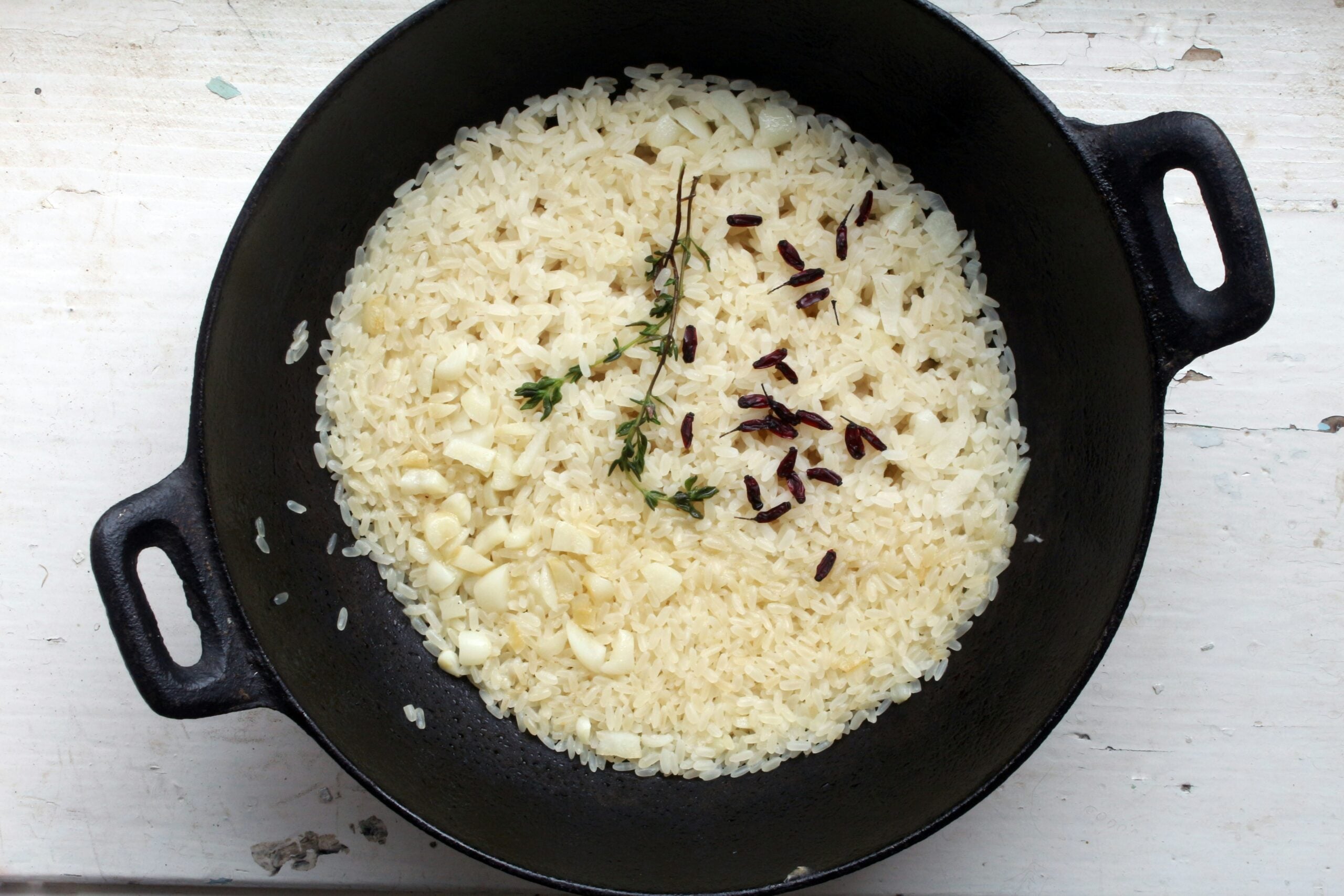 A black pot filled with uncooked white rice, garlic pieces, fresh thyme sprigs, and a few clove buds, sitting on a white surface.