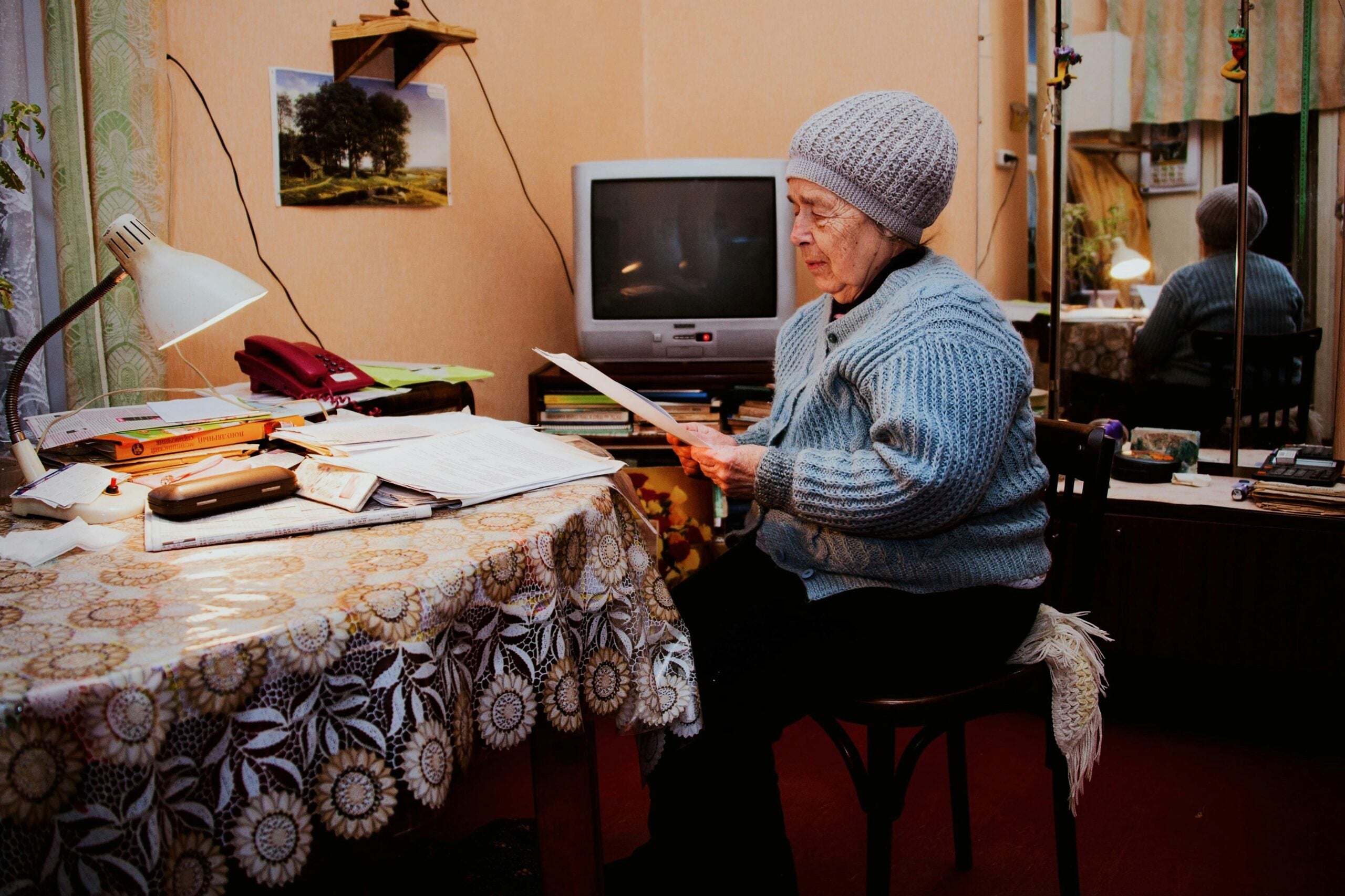 An elderly woman wearing a knit hat and blue sweater sits at a table covered with papers, carefully reading a document about her retirement savings in a cozy room with a TV, lamp, books, and a mirror reflecting part of the scene.