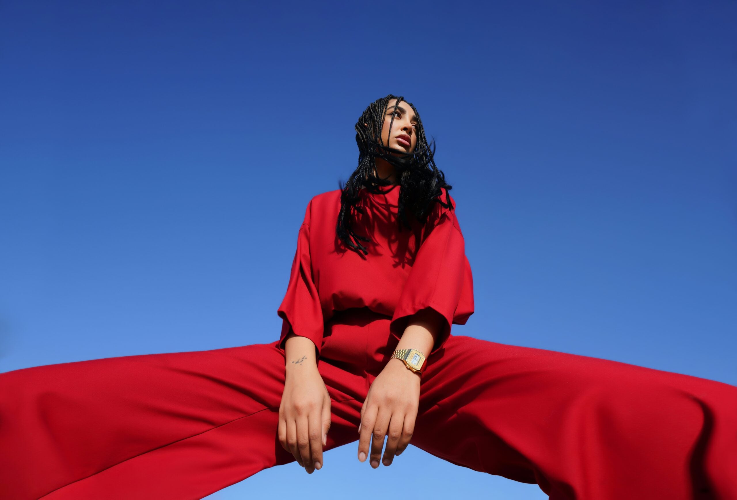 Person in a bold red outfit poses with legs apart, hands resting between knees, against a clear blue sky, creating a striking color contrast and dramatic low-angle perspective.