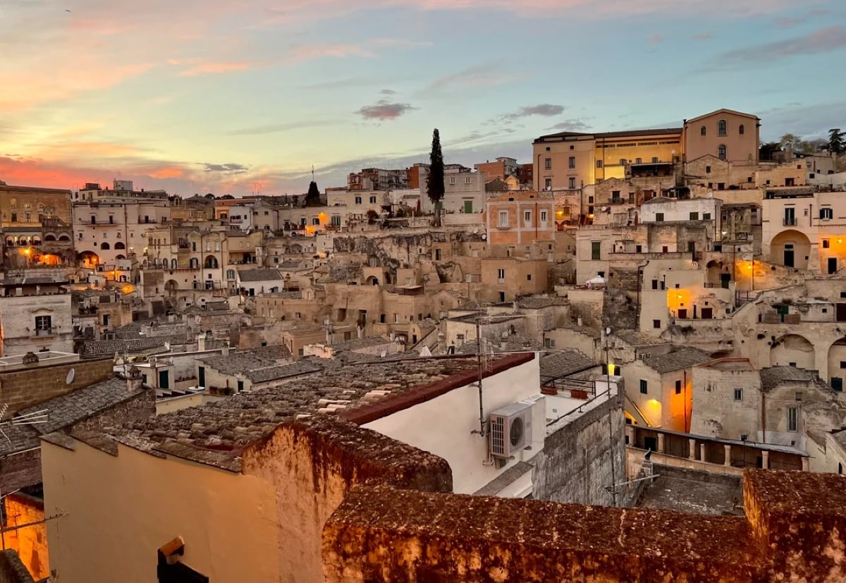 Ancient stone buildings with flat roofs crowd a hillside at sunset. Warm yellow lights glow from windows, and the sky is streaked with pink and blue. The scene appears calm and historic.
