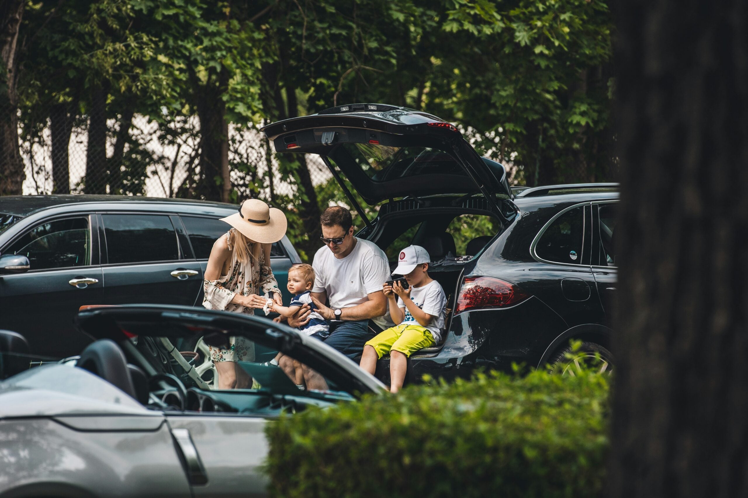 A family of four stands near an open car trunk in a parking lot. The parents look at their phones, one child sits in the trunk, and another child looks at a device. Other cars and trees are in the background.