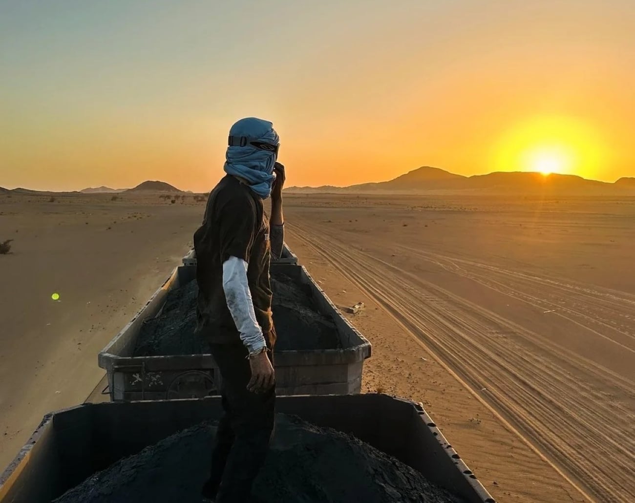A person stands atop a freight train car loaded with minerals, wearing a headscarf and looking at the desert landscape and mountains as the sun sets, casting an orange glow over the scene.