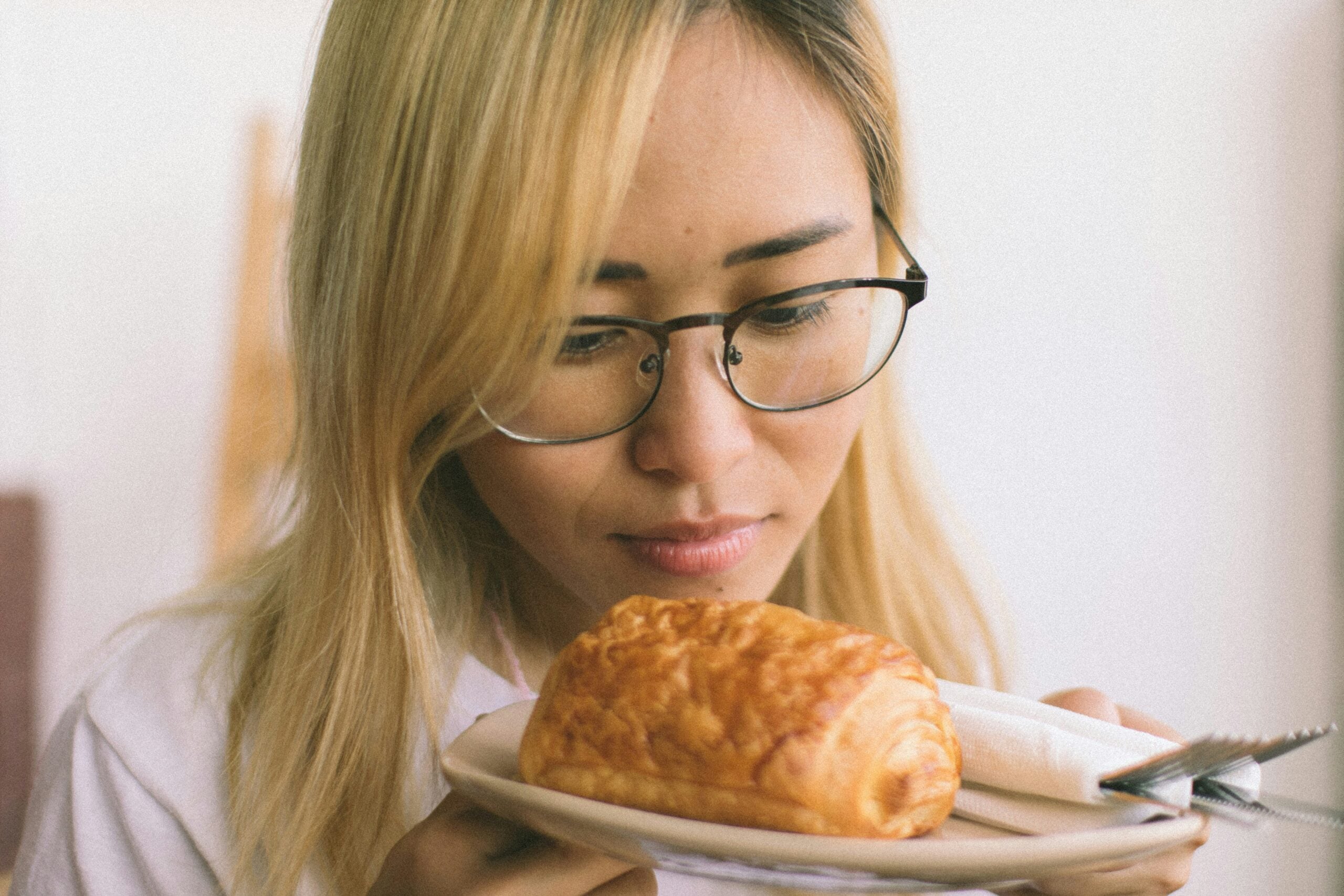 A woman with blonde hair and glasses looks closely at a pastry on a plate, which also holds a folded napkin and a fork.