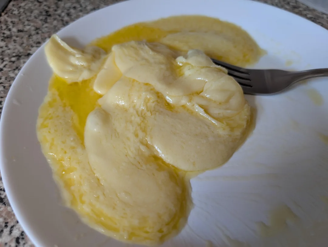 Melted cheese with a pool of oil on a white plate, next to a fork on a speckled countertop.