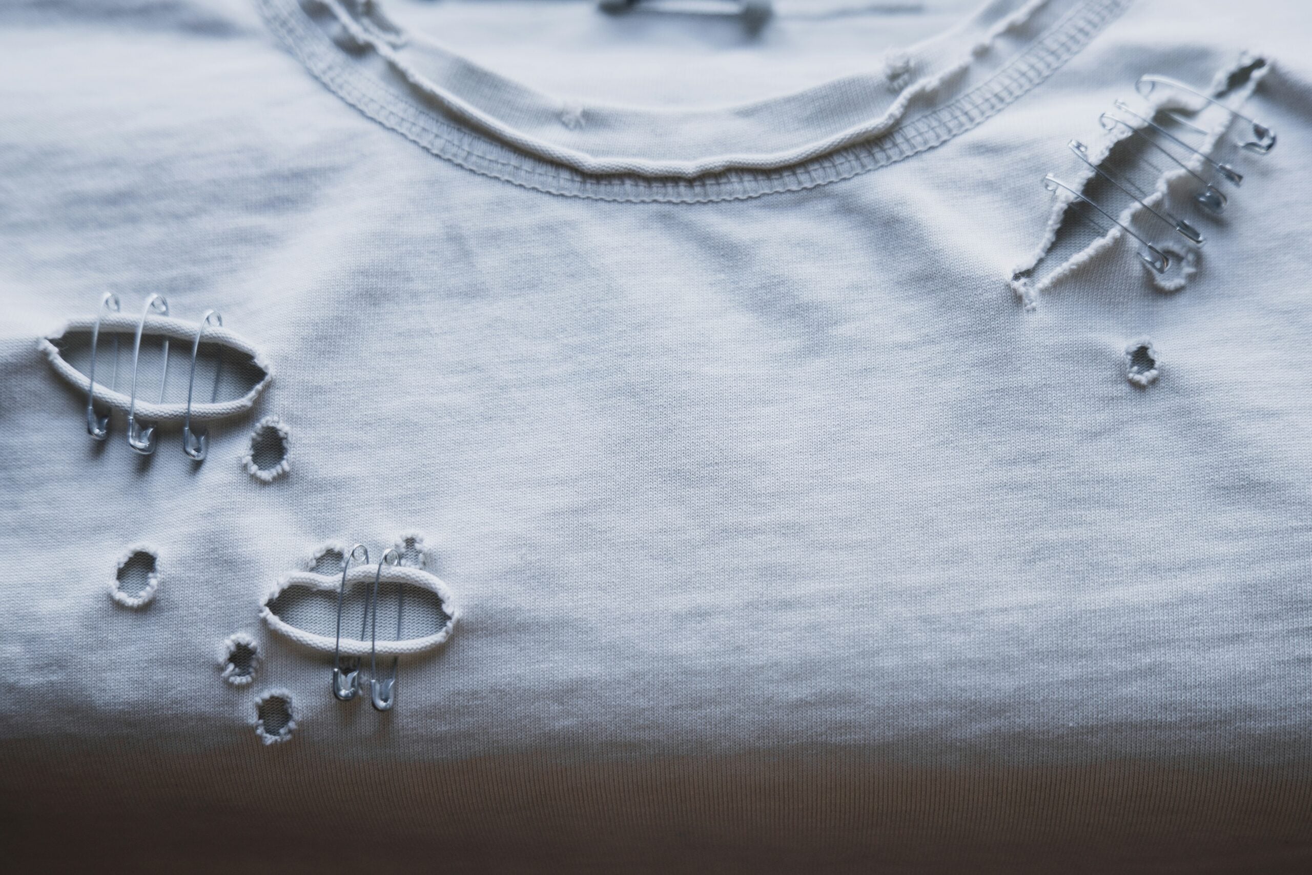 A close-up of a white t-shirt with intentional distressed holes and visible white stitching, creating a decorative, worn-out look on the fabric near the collar.