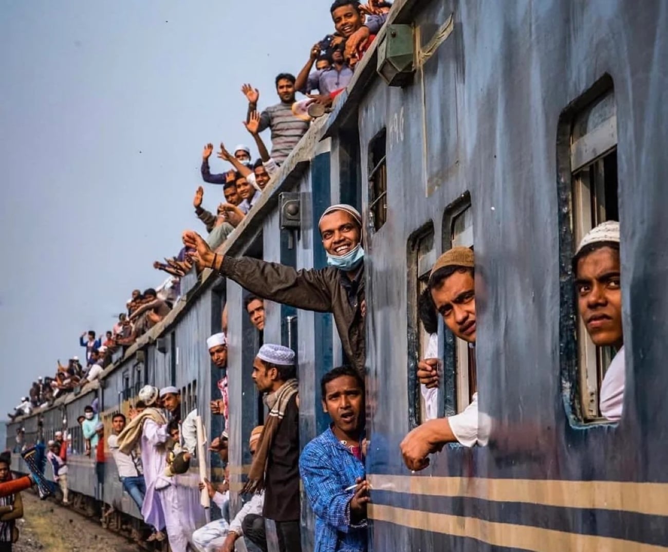 Many people crowd on and inside a moving blue train, some waving and smiling from windows and the roof. The atmosphere is lively and joyful under a clear sky.