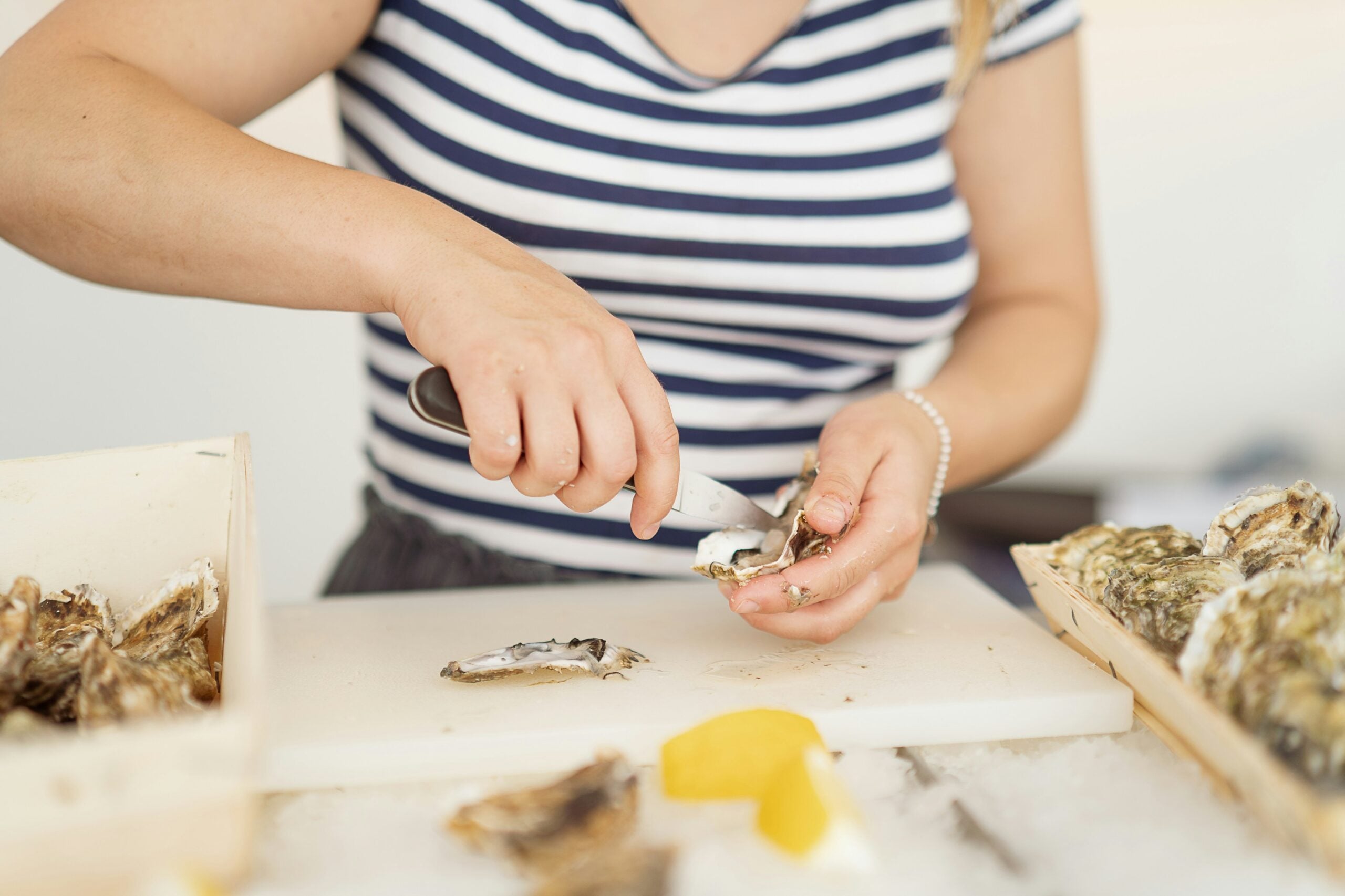A person in a striped shirt uses an oyster knife to open an oyster on a white cutting board, with lemon wedges and more oysters nearby.