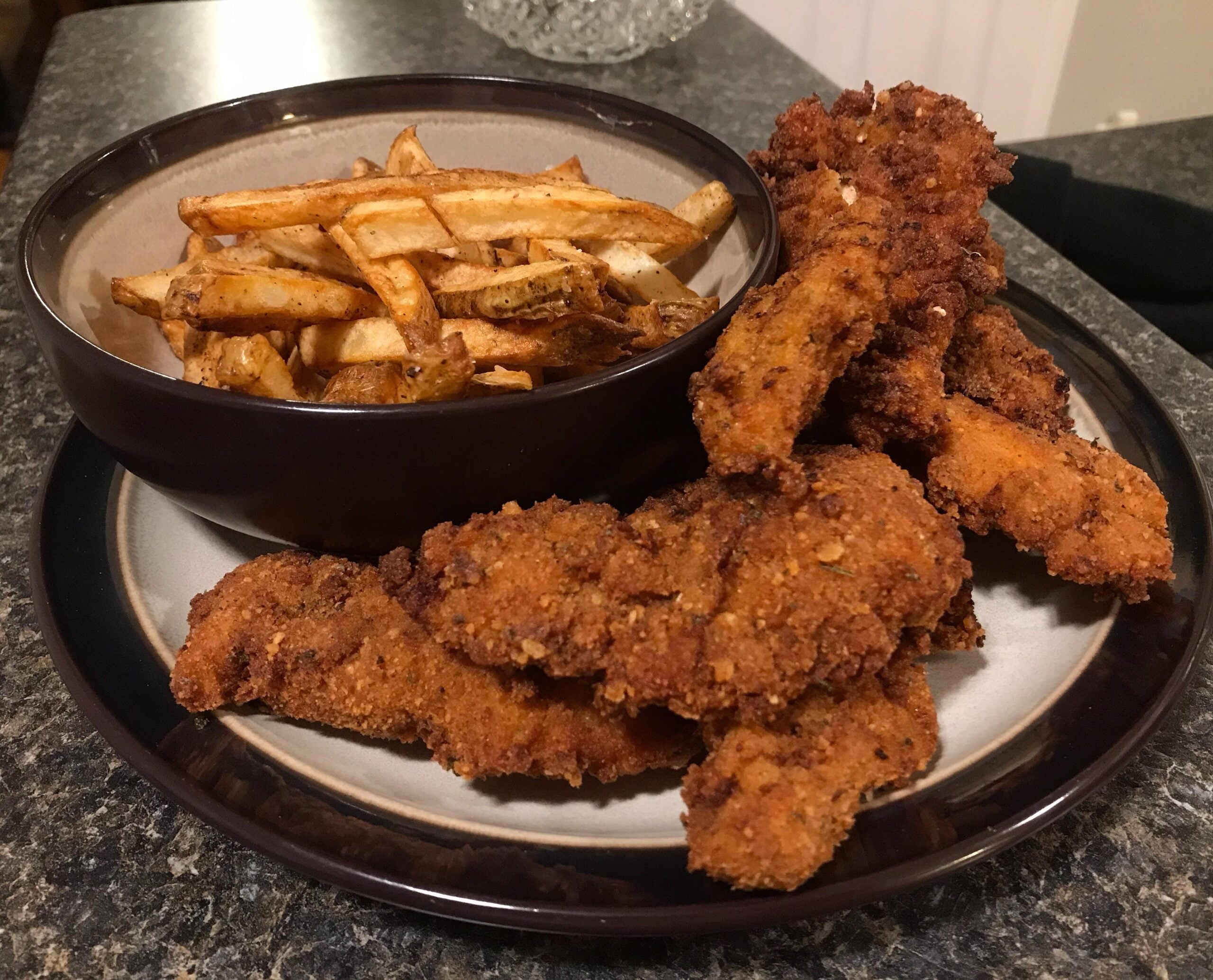 A plate with crispy fried chicken tenders and a bowl of golden-brown French fries on a dark countertop.