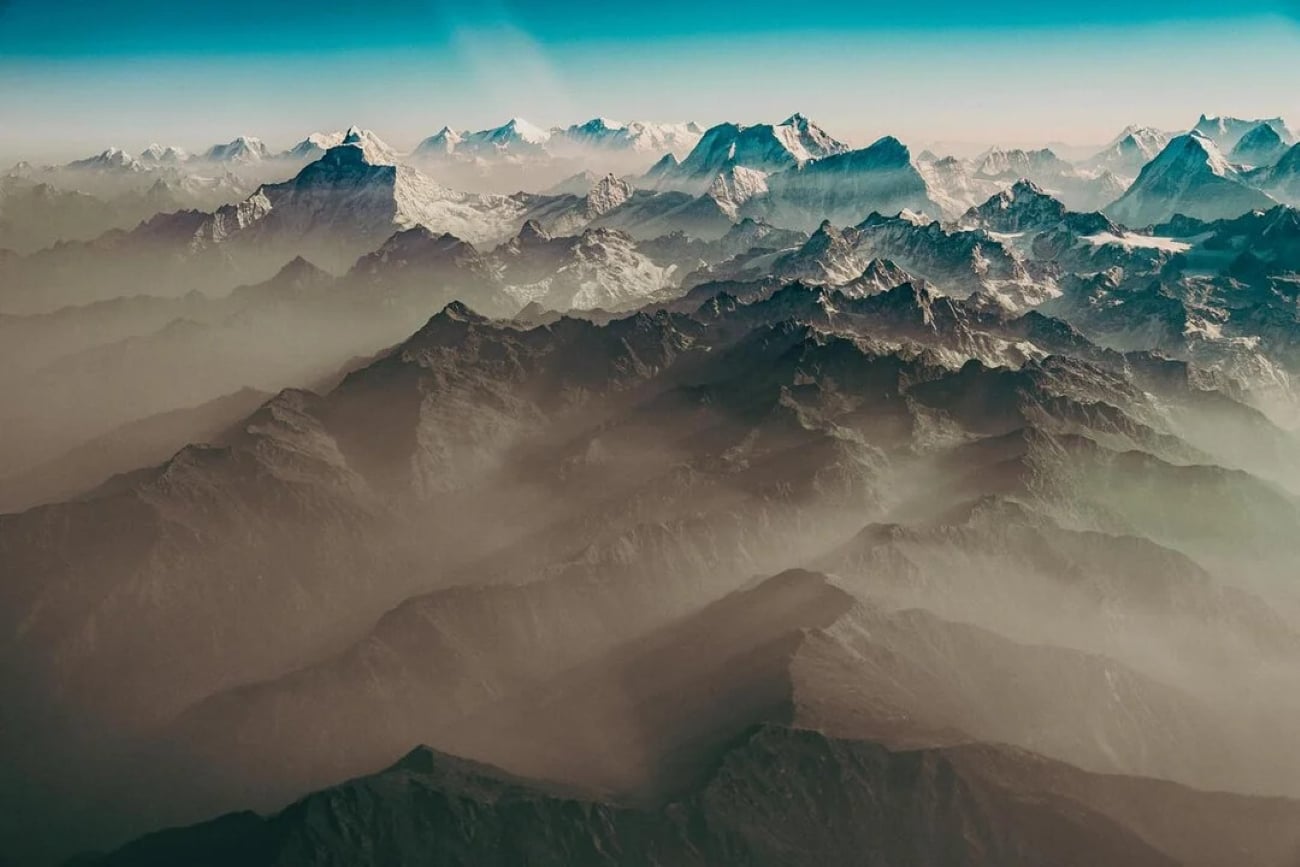 Aerial view of a vast mountain range with jagged, snow-capped peaks under a clear blue sky, as seen through airplane windows. The valleys below are partially shrouded in mist, creating layers of shadow and light across the landscape.