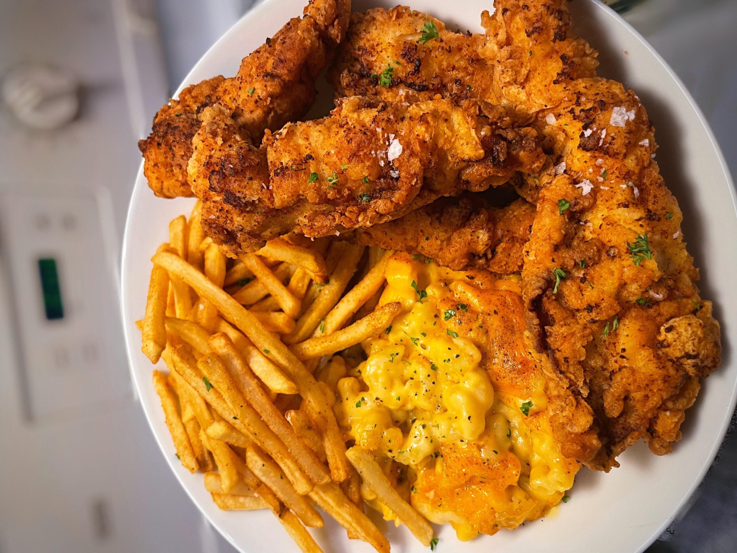 A plate of crispy fried chicken tenders, golden French fries, and creamy macaroni and cheese, garnished with herbs.