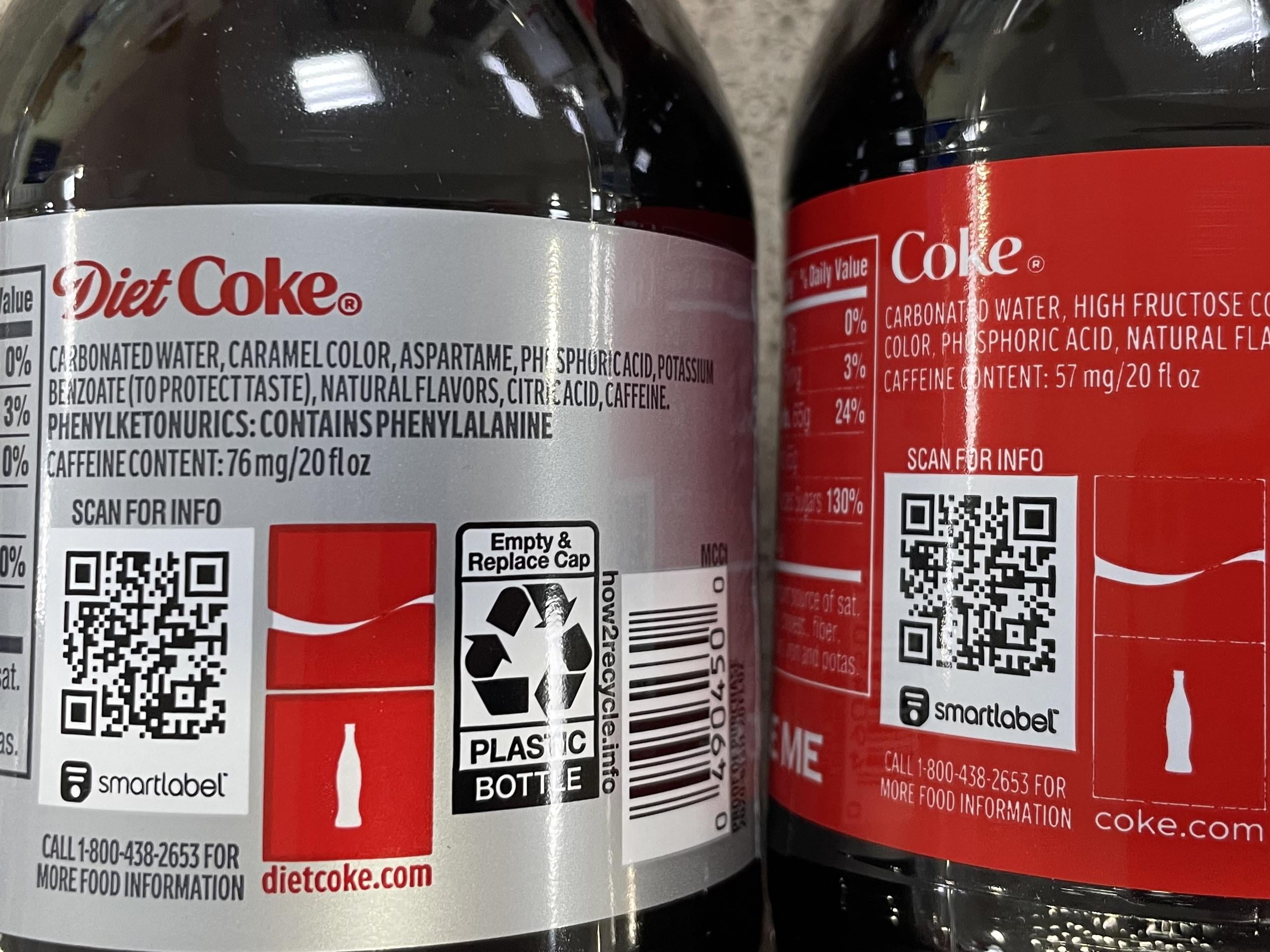 Close-up of two soda bottles showing Diet Coke and Coca-Cola labels, including ingredients, nutritional facts, QR codes, and recycling information. Labels emphasize artificial sweeteners and caffeine content in Diet Coke.