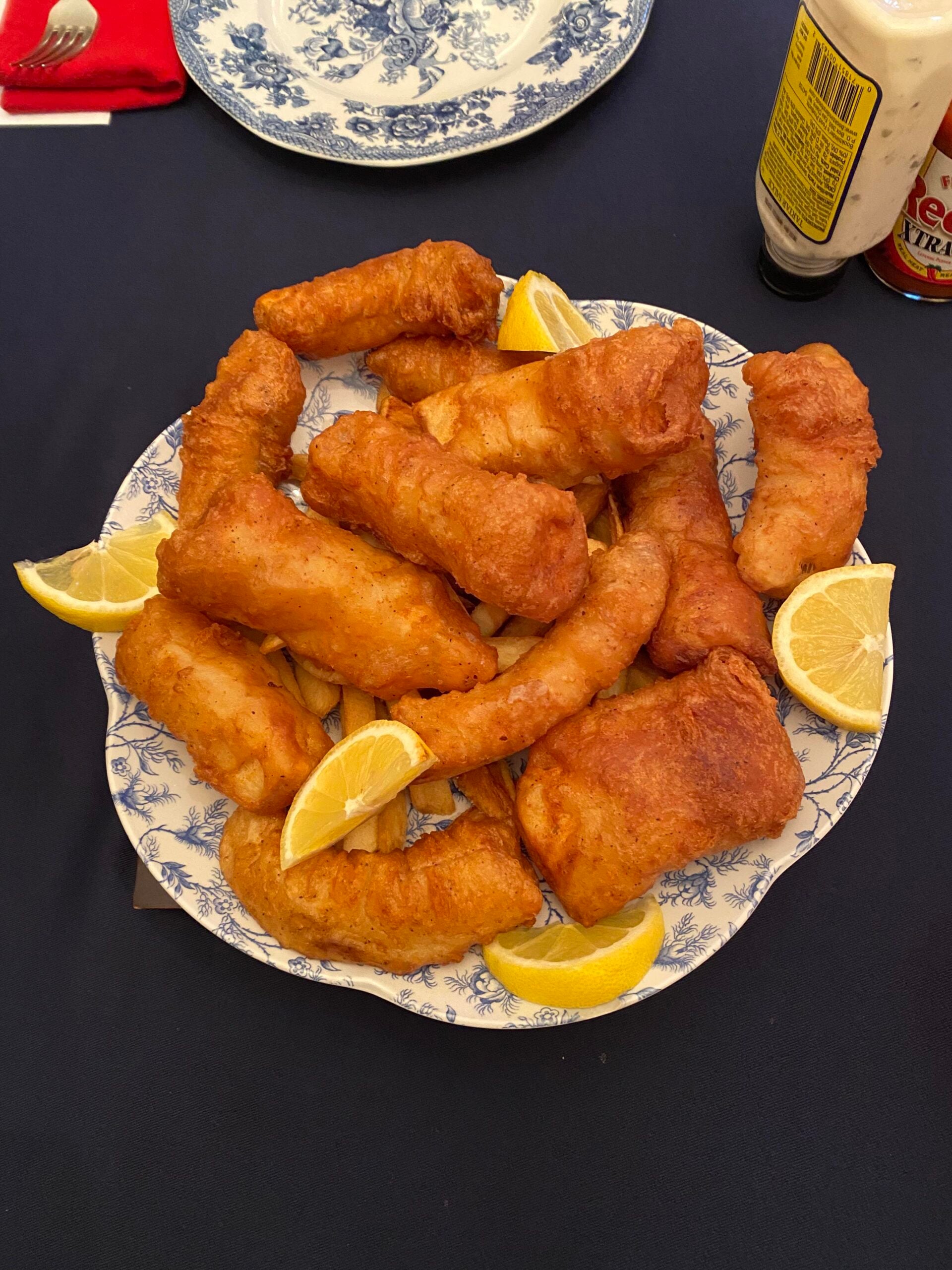 A blue and white plate holds pieces of fried fish and lemon wedges, set on a dark tablecloth with a bottle of tartar sauce and a partially visible decorative plate nearby.