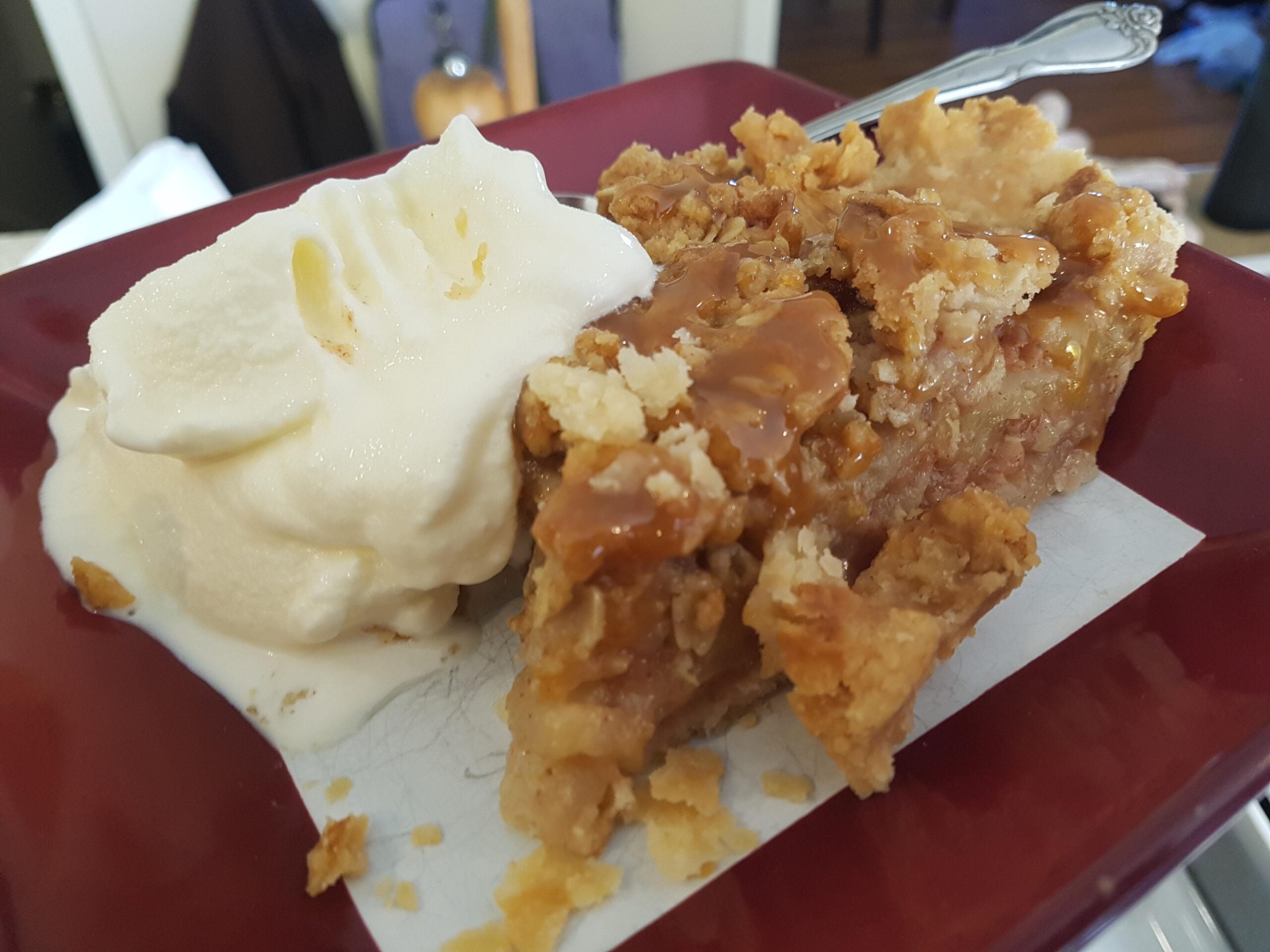 A slice of apple pie with crumb topping and caramel sauce served on a red plate with a scoop of vanilla ice cream and a spoon.