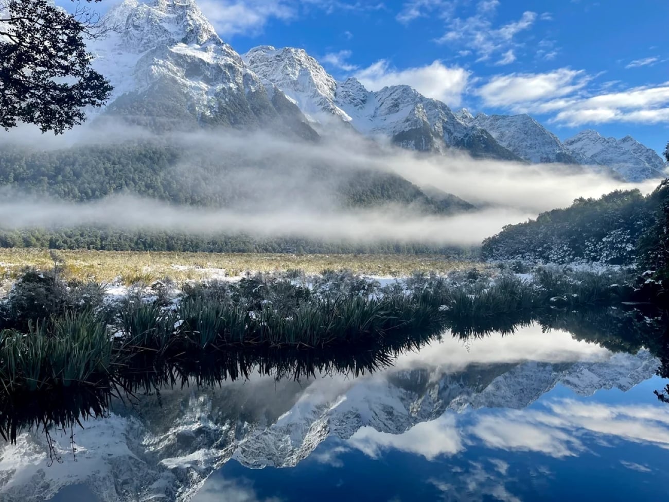 Snow-capped mountains with a layer of mist, lush green trees, and clear water below reflecting the landscape and blue sky with scattered clouds.