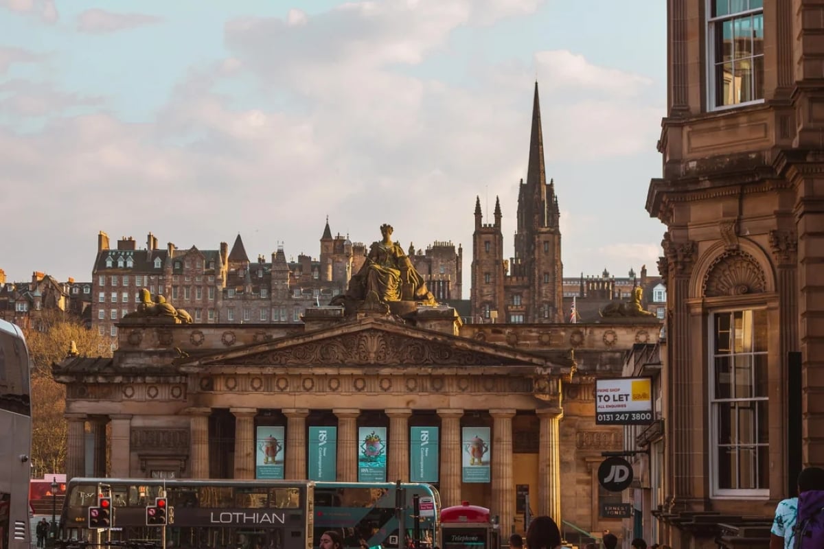 Historic building with statues on its roof, surrounded by classic architecture. Buses and cars pass by, with a cityscape and church spires in the background under a partly cloudy sky.