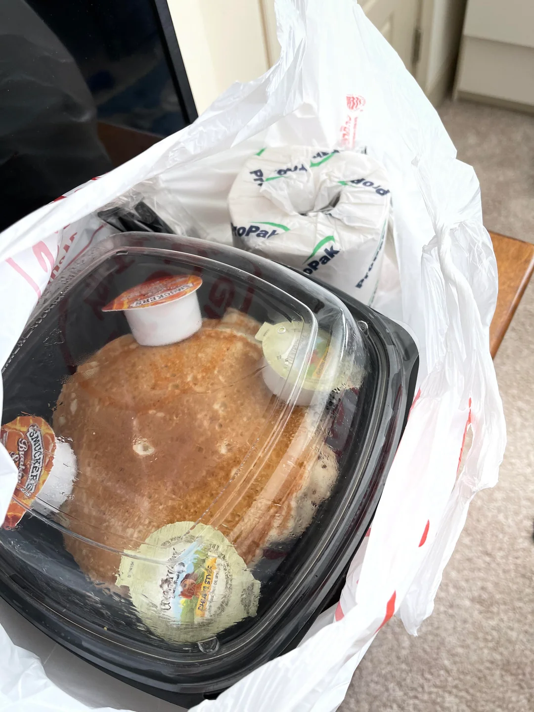 A takeout container with pancakes, butter, and syrup sits inside a plastic bag next to wrapped utensils, on a carpeted floor near a wooden table.