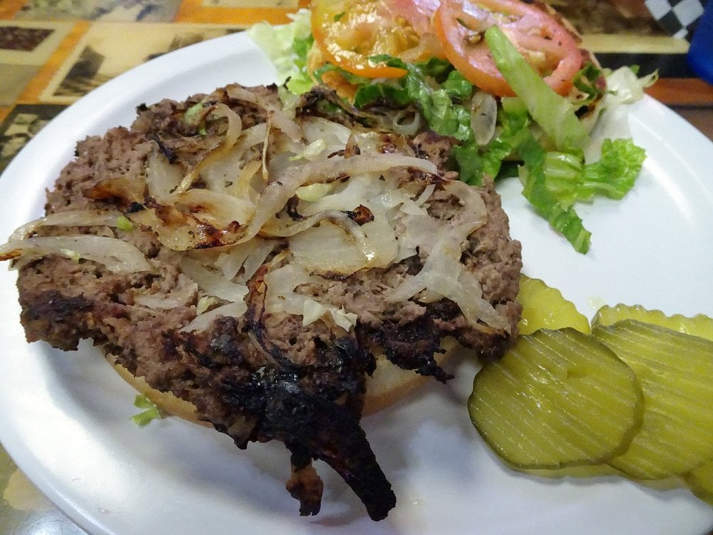 Open-faced hamburger patty topped with grilled onions, served on a white plate with sliced pickles and a side salad of lettuce and tomatoes.