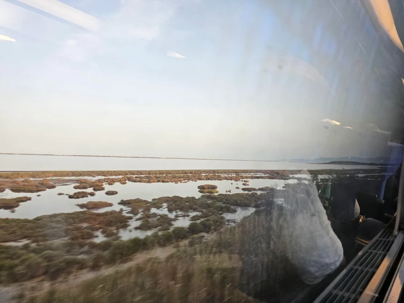 View from a train window showing a coastal marshland with low water, patches of vegetation, and a blue sky. Reflections of passengers and seats are visible on the glass.