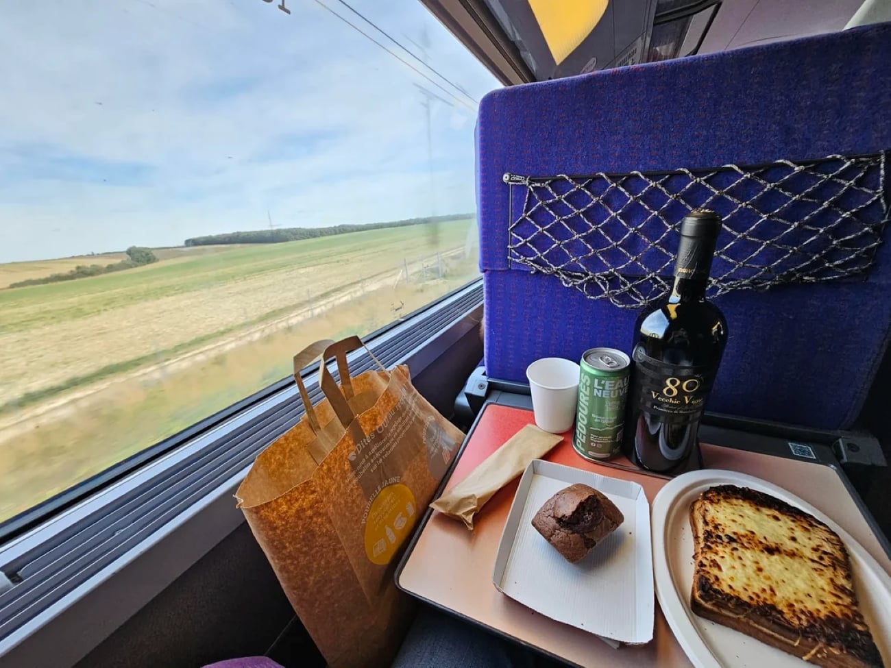 A meal with lasagna, bread, a muffin, wine, and a can is set on a tray table by a train window showing fields and blue sky. A paper bag sits beside the food. The seat has a blue cover with a storage net.