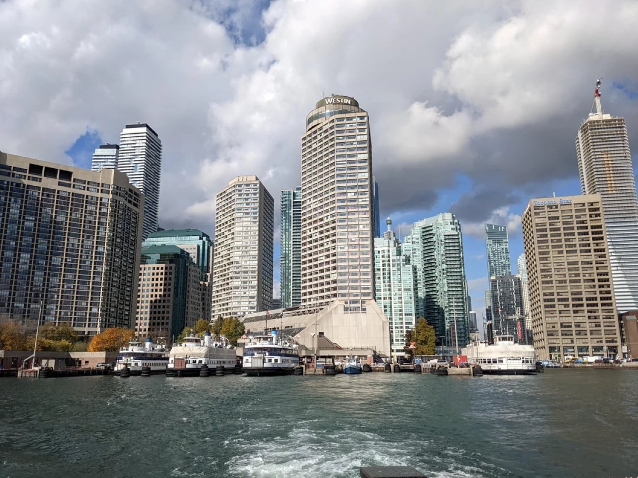 A city skyline with tall modern buildings next to a waterfront, boats docked along the shore, and a partly cloudy sky overhead.