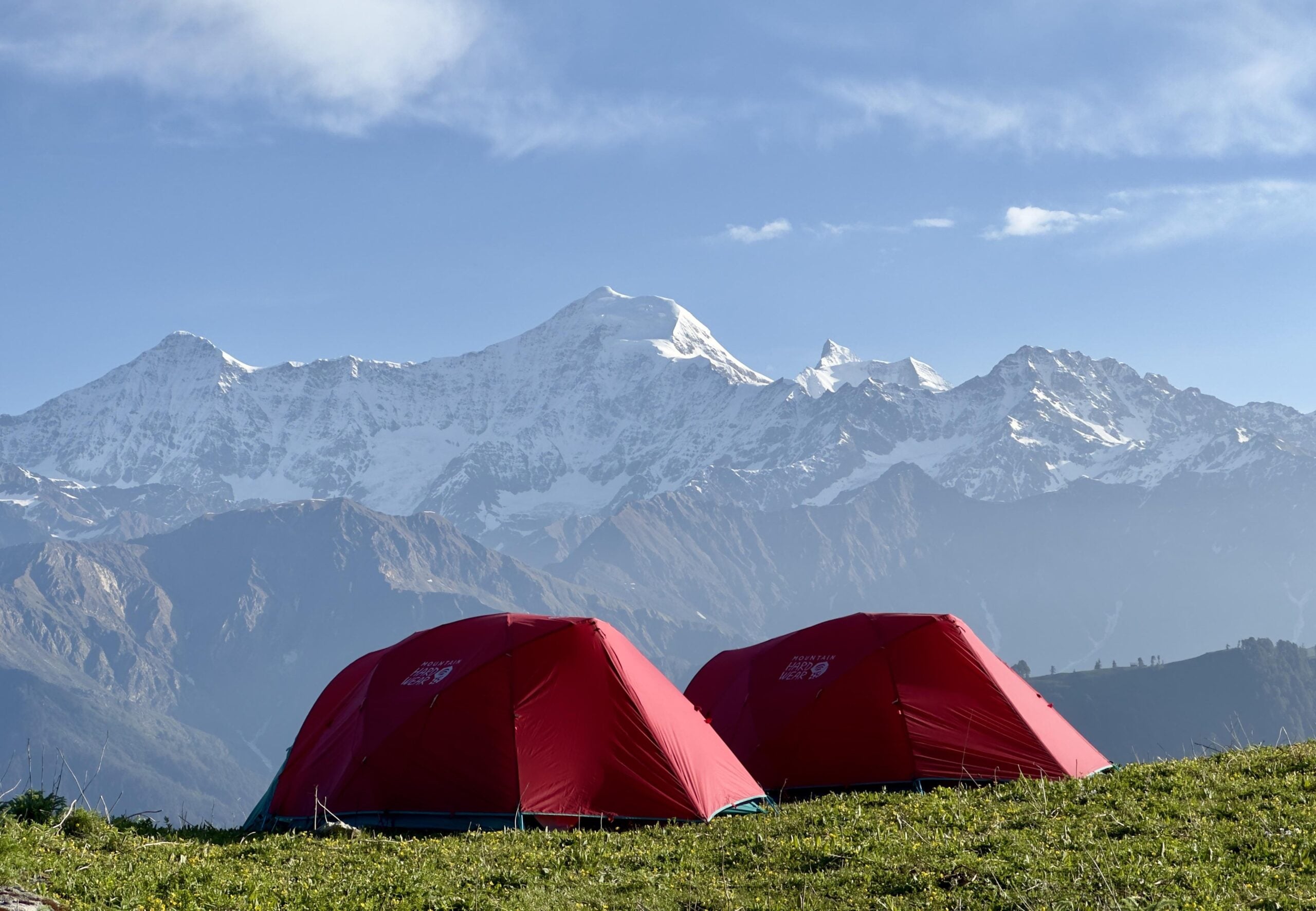 Two red tents sit on grassy ground with a backdrop of snow-capped mountains and a blue sky. Sunlight highlights the tents and the landscape, creating a serene outdoor camping scene.