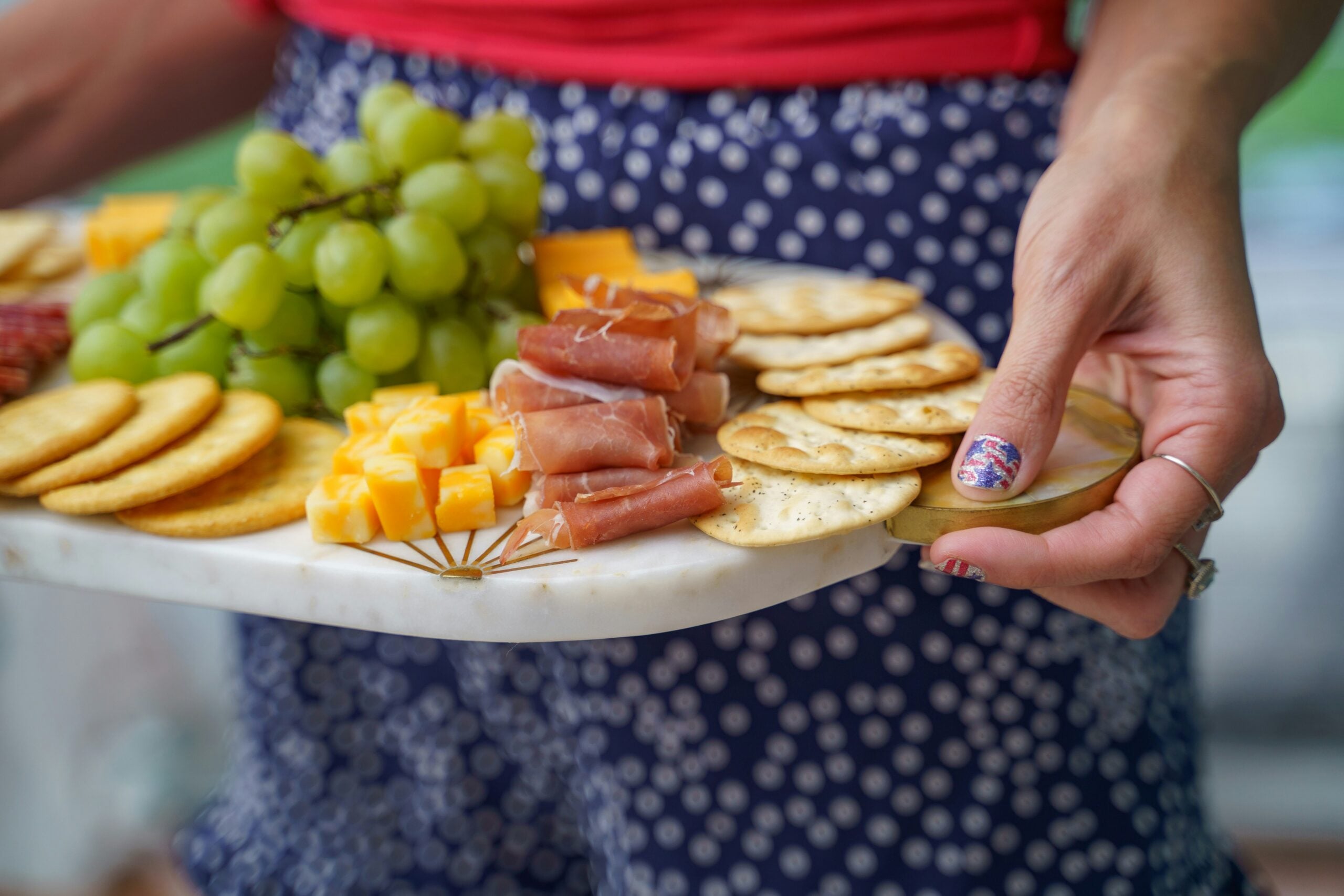 A person holds a marble tray with green grapes, cheese cubes, crackers, and prosciutto. The person wears polka dot shorts and has decorative nails.