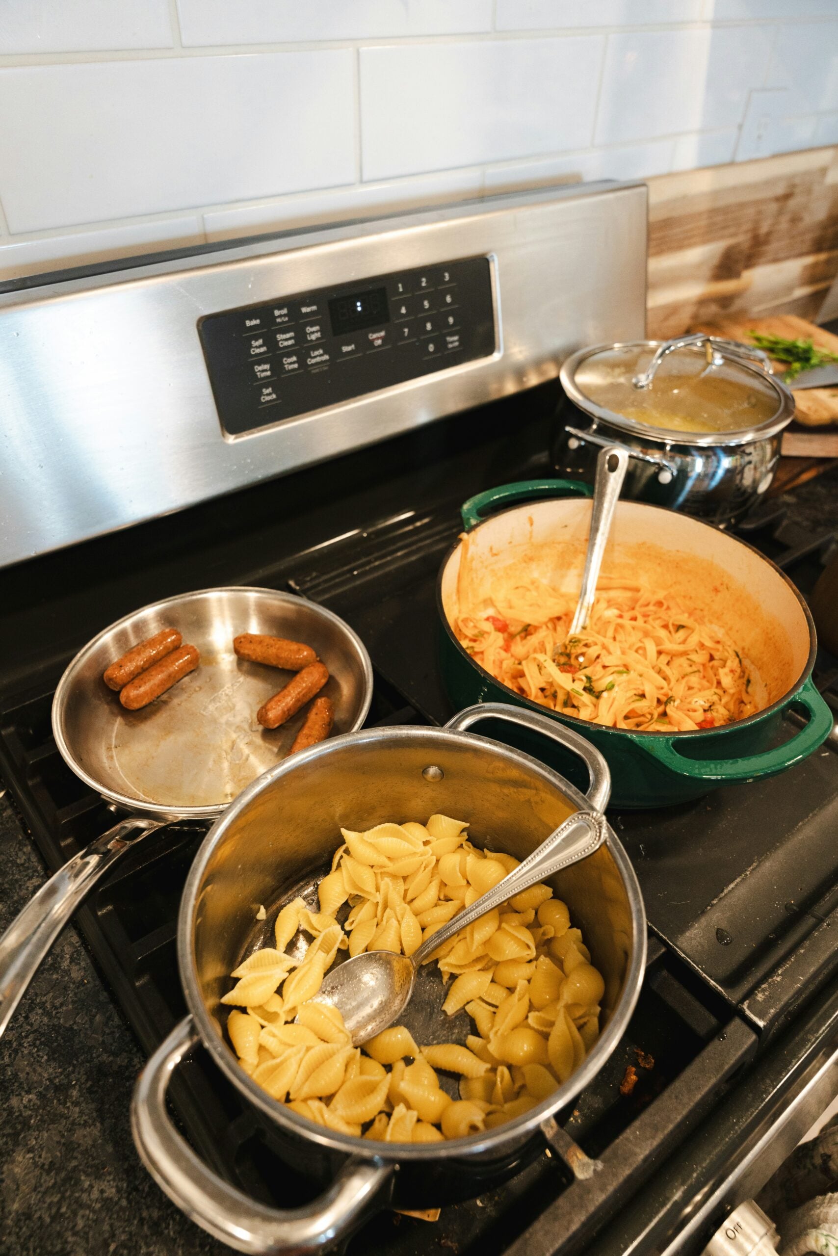Four-burner stove with a pot of shell pasta, a pan with sausages, a pot with boiling water, and a Dutch oven containing pasta with a creamy sauce, all cooking at once.
