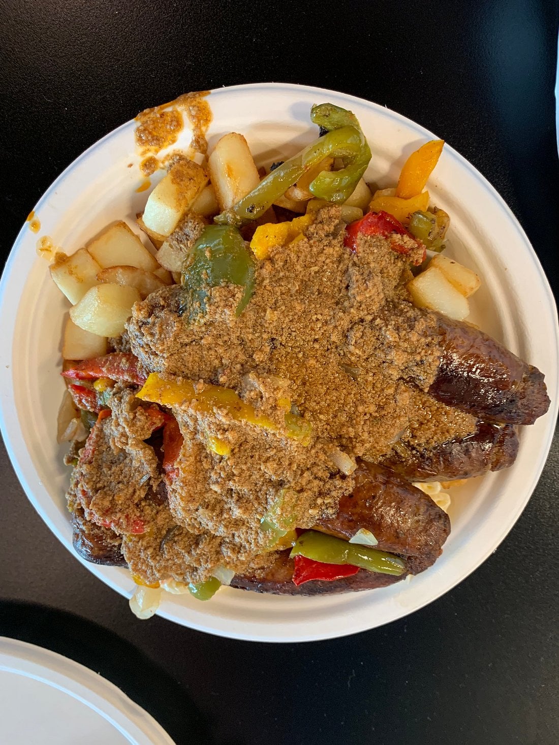 A white plate filled with sausage links, diced potatoes, sautéed bell peppers, onions, and a brown crumbly topping, all served on a dark table.