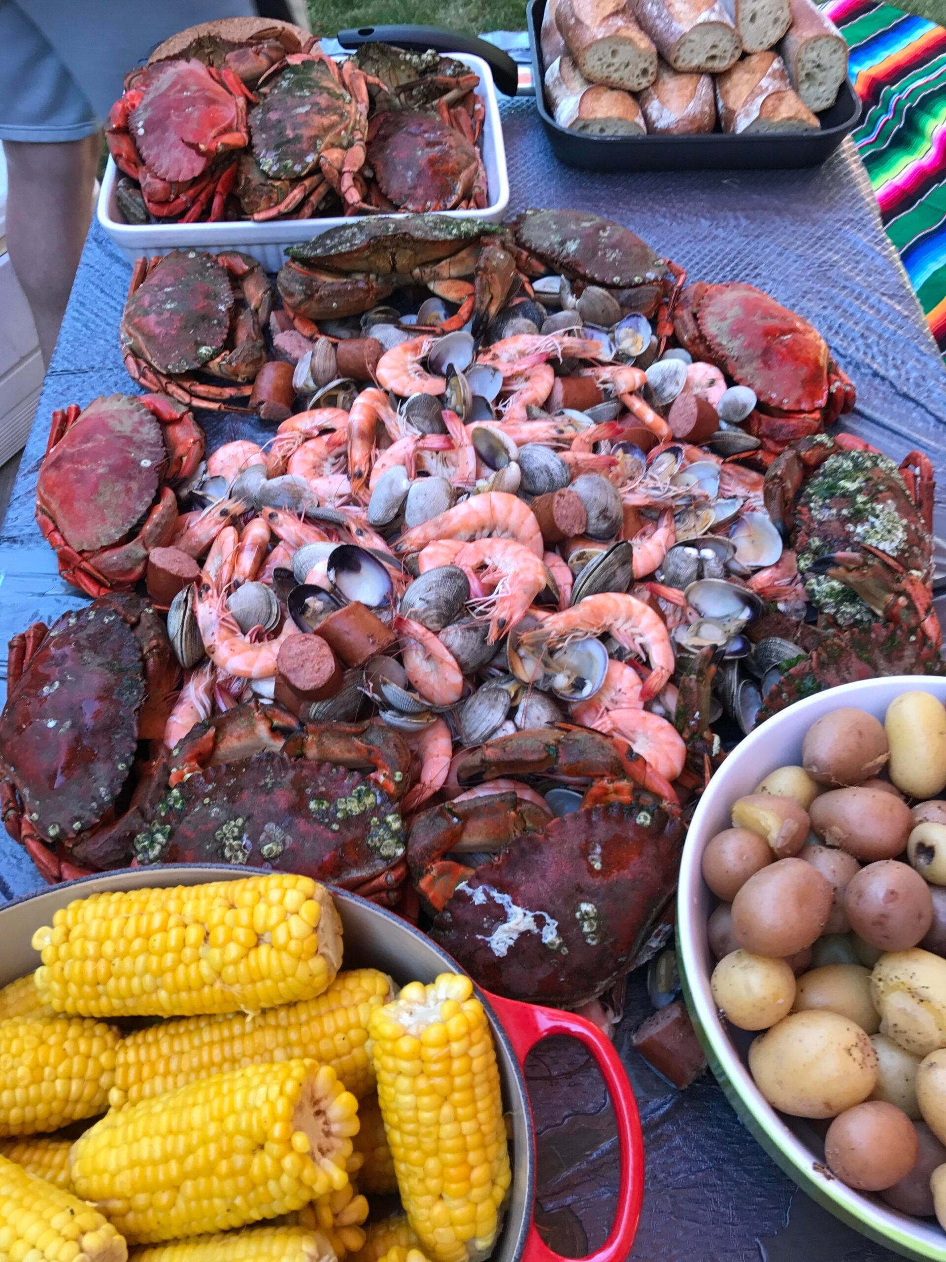 A table covered with cooked crabs, shrimp, and clams, surrounded by bowls of corn on the cob, potatoes, and bread slices, ready for a seafood feast.