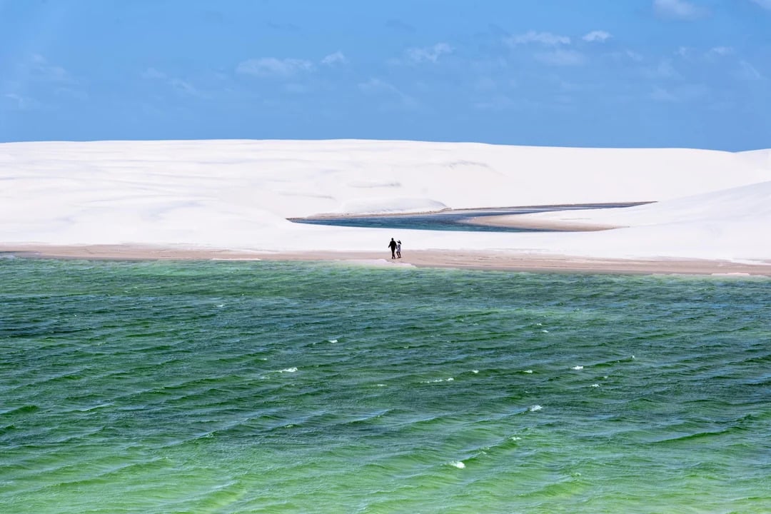 Two people walk along a sandy stretch between green water and white sand dunes under a bright blue sky with scattered clouds.