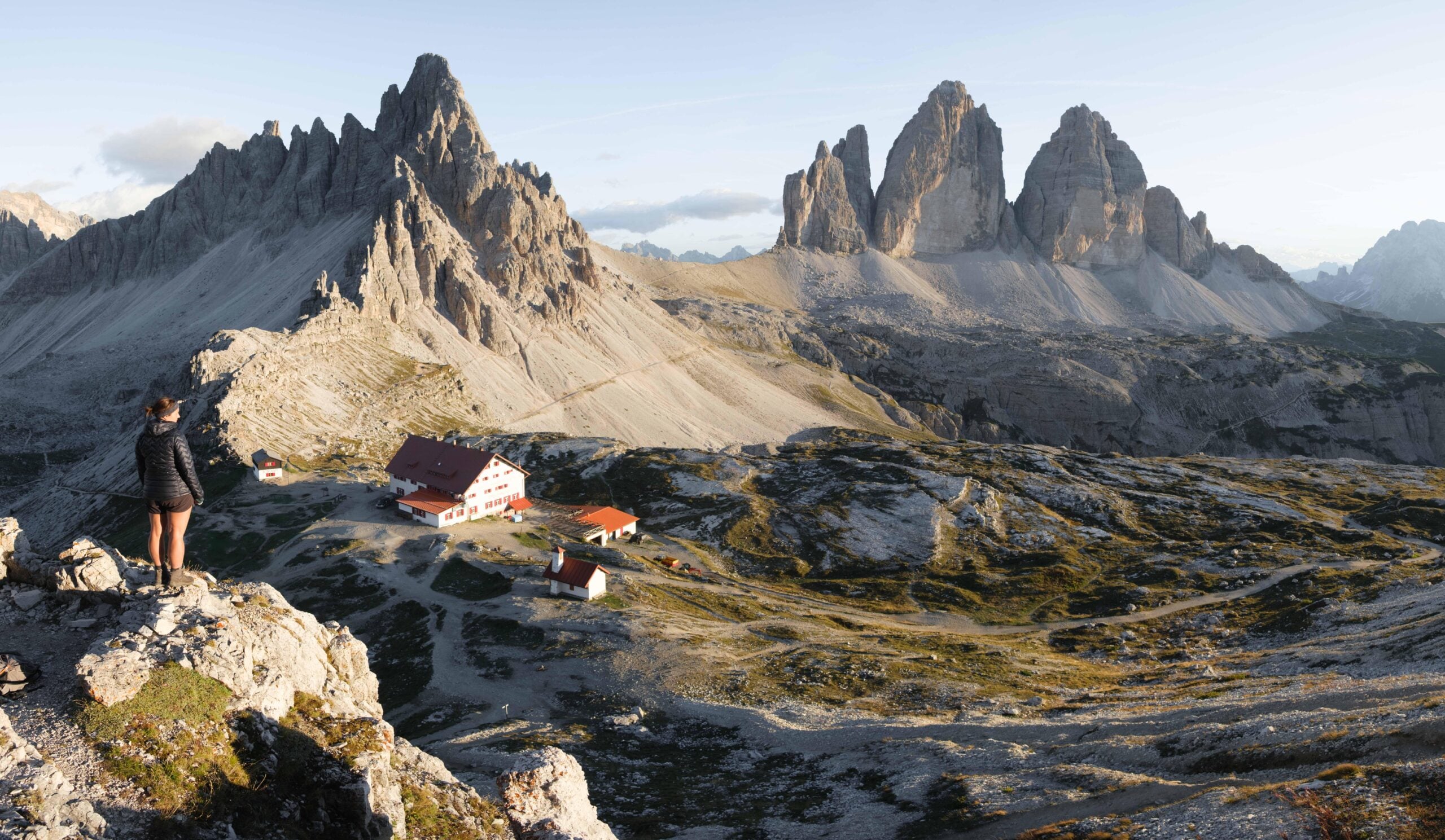 A person stands on a rocky outcrop overlooking a mountain hut and cabins, with sharp, dramatic peaks rising in the background under a clear sky.