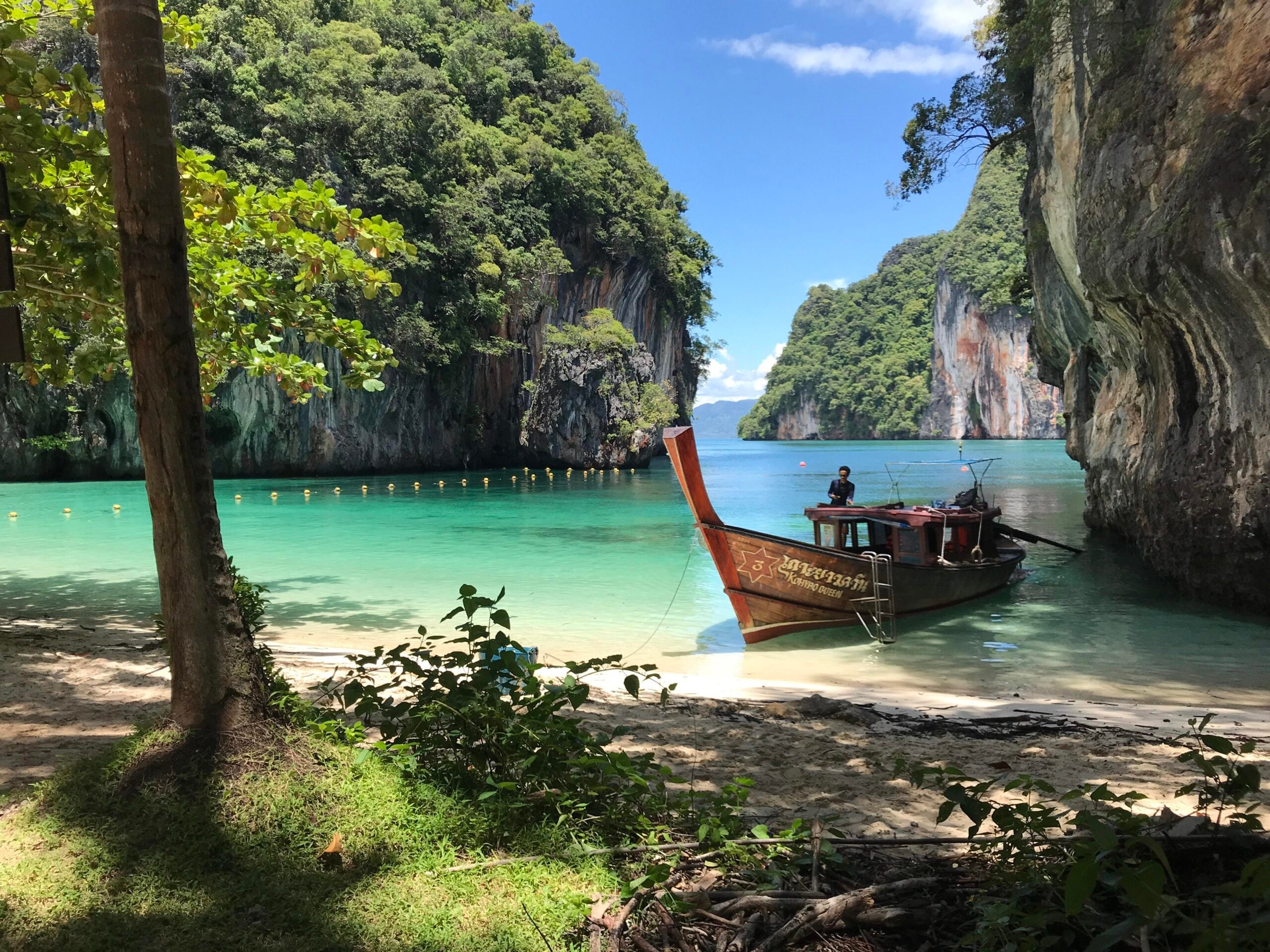 A traditional wooden boat is anchored in clear turquoise water between lush, rocky cliffs under a bright blue sky, surrounded by trees and a sandy shore in the foreground—an idyllic scene set near some of the region’s most incredible beaches.