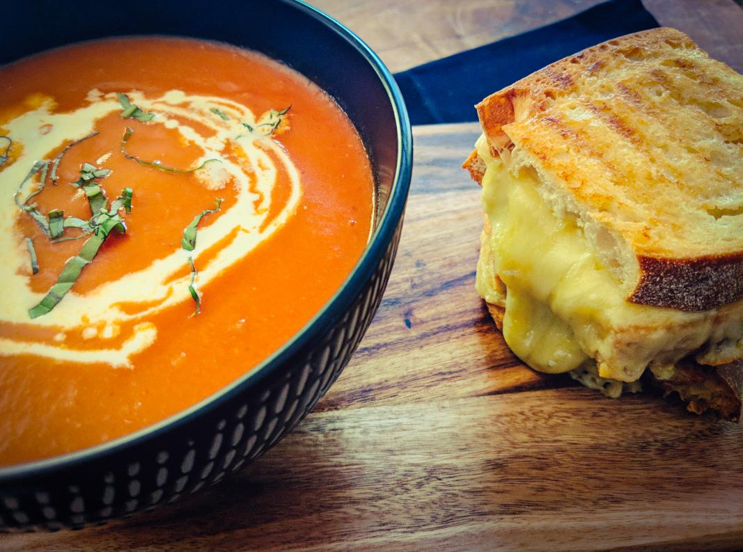 A bowl of tomato soup with cream and herbs sits next to a classic grilled cheese sandwich on a wooden board, creating the perfect pairing for fans of grilled cheese sandwiches.