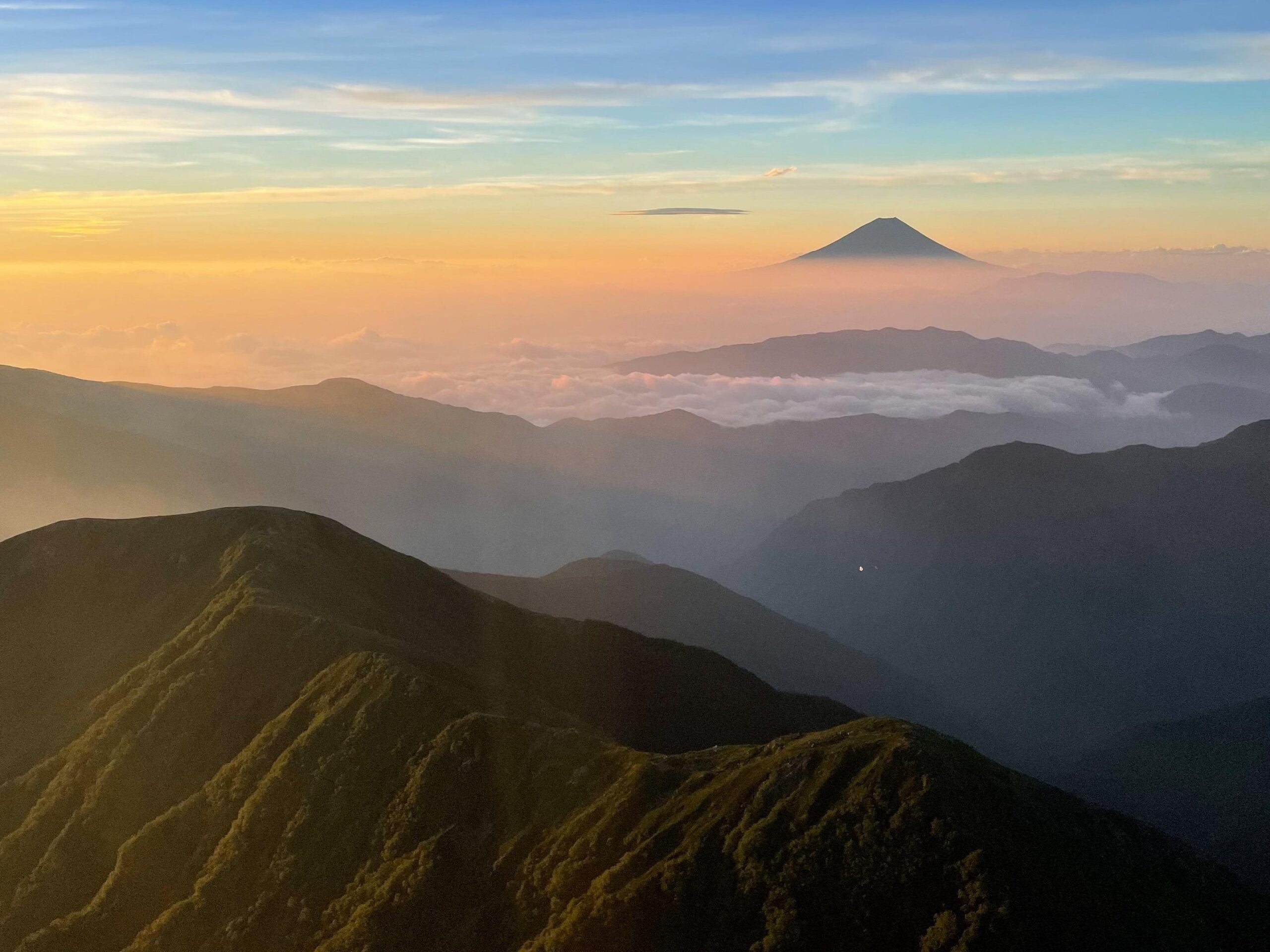Distant mountain peaks with lush greenery at sunrise, with a layer of clouds in the valley and a tall, conical volcano on the horizon under a colorful sky.