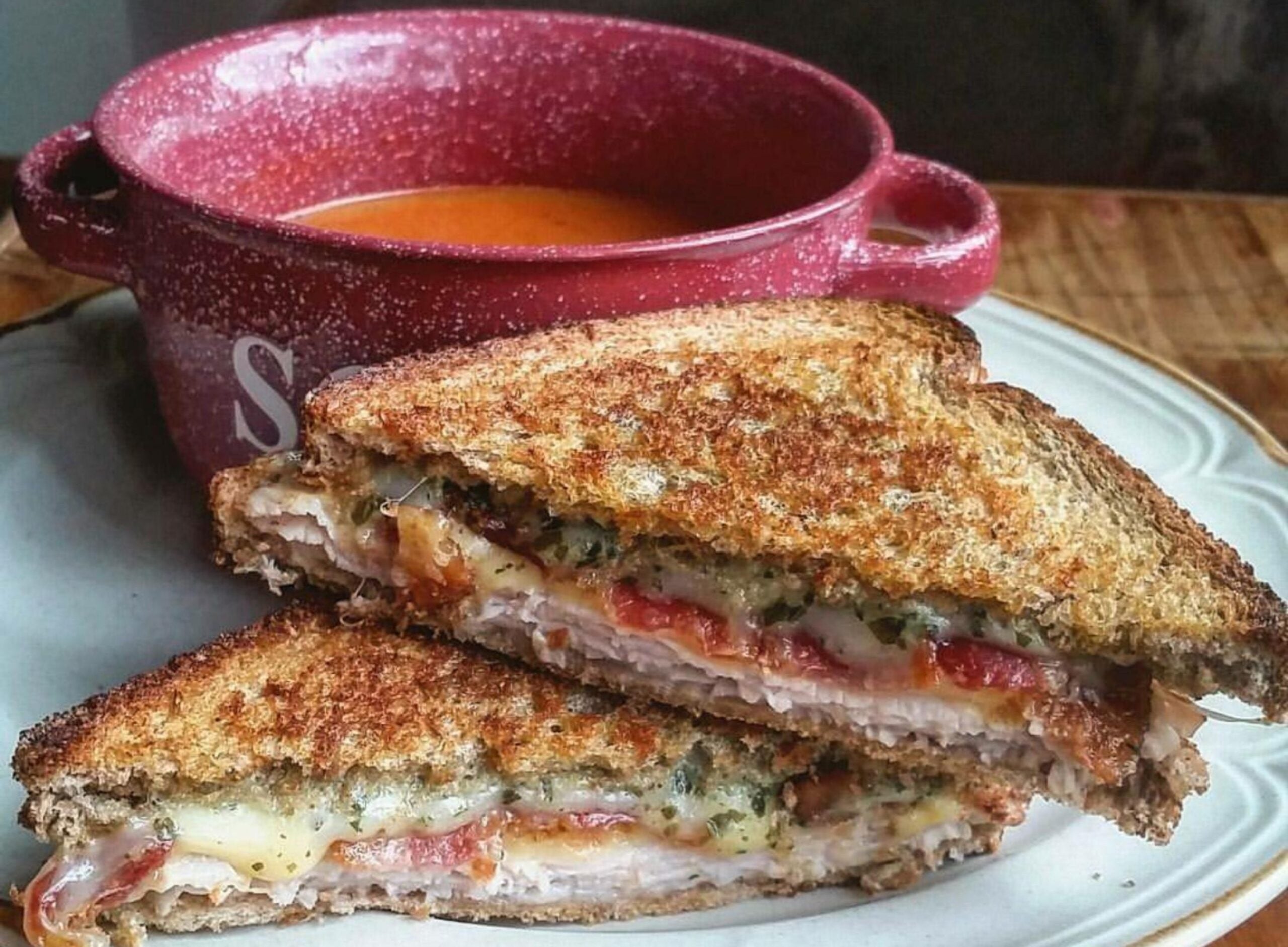 A grilled sandwich with melted cheese and turkey is cut in half on a plate, next to a red bowl filled with tomato soup.