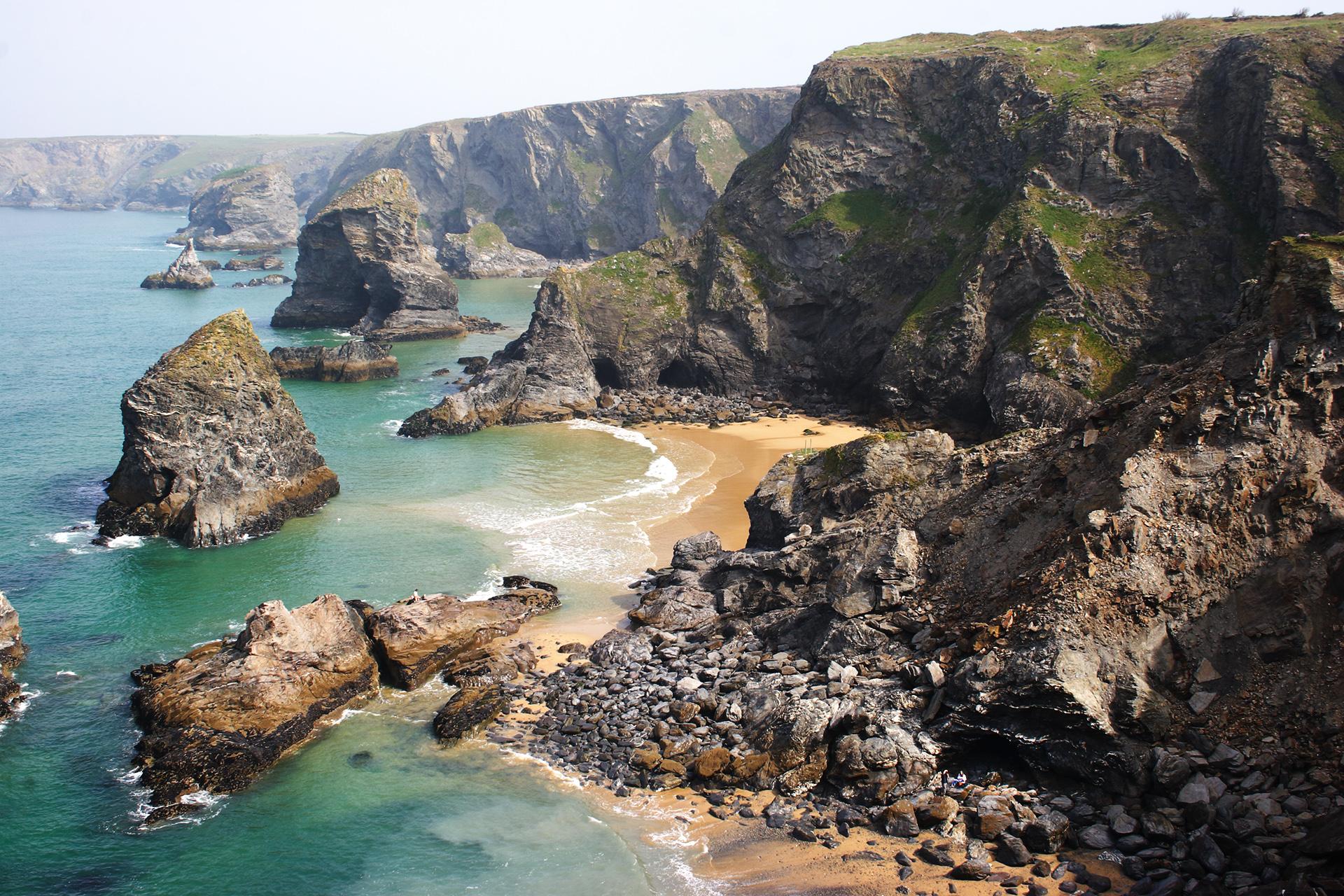Rocky cliffs and sea stacks surround sandy coves along a clear turquoise shoreline, with waves gently hitting the beach. The landscape looks rugged and natural under a partly cloudy sky.