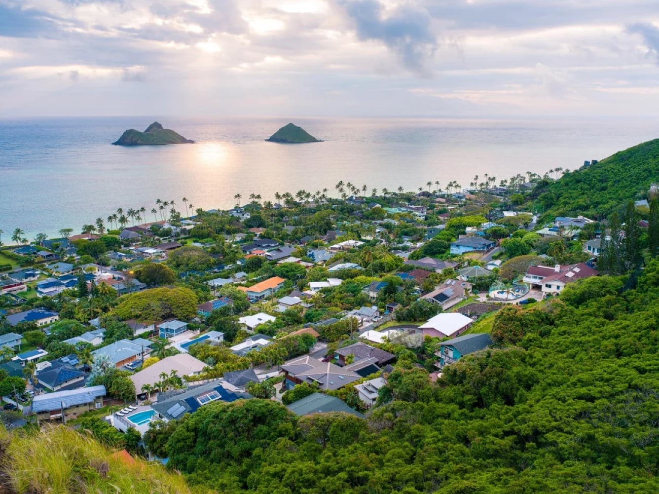A coastal town with lush greenery, houses, palm trees, and two small islands offshore under a cloudy sky at sunset—an idyllic scene often found in tropical locations.