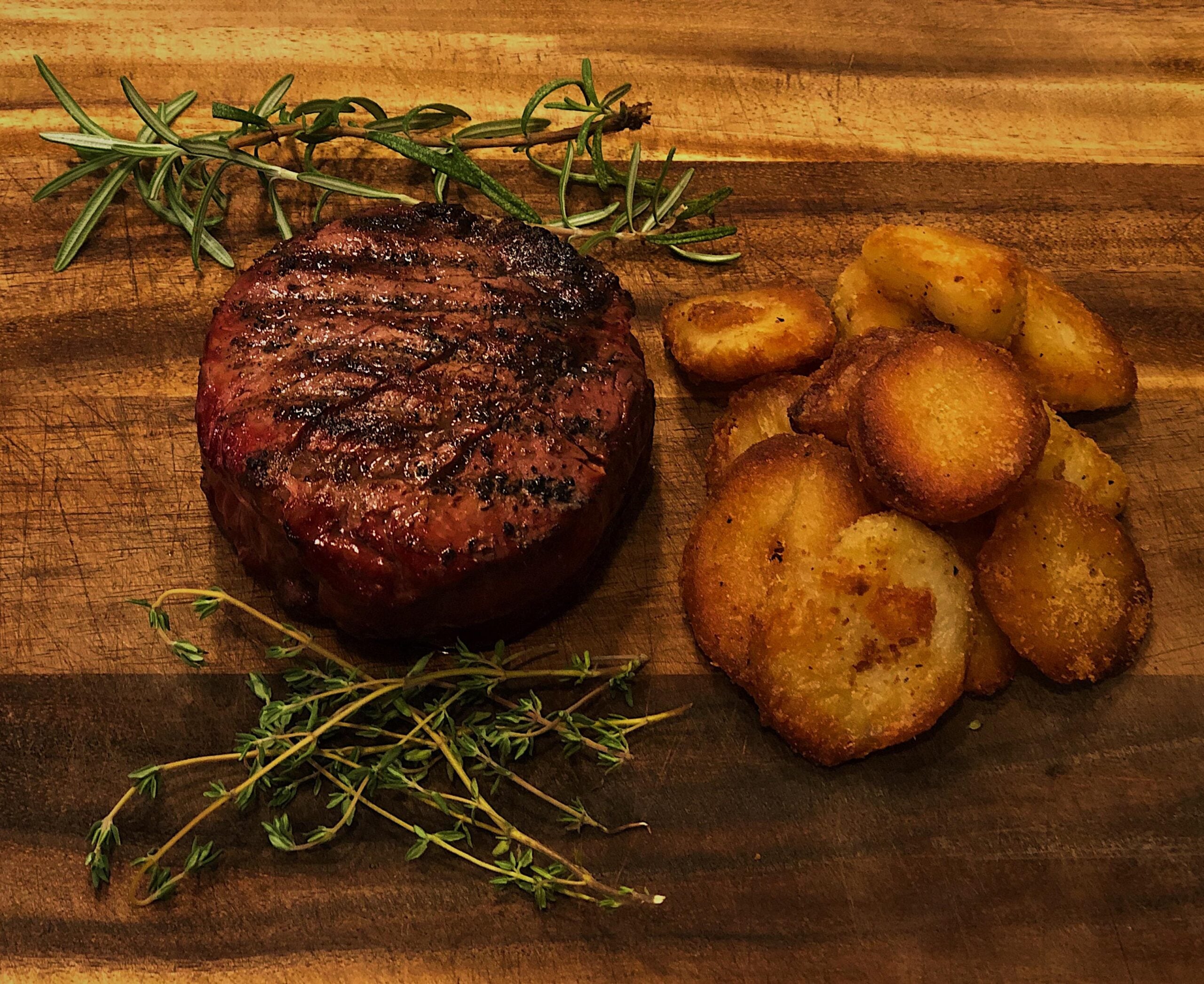Grilled steak with grill marks sits beside golden roasted potatoes and fresh rosemary and thyme on a wooden board.
