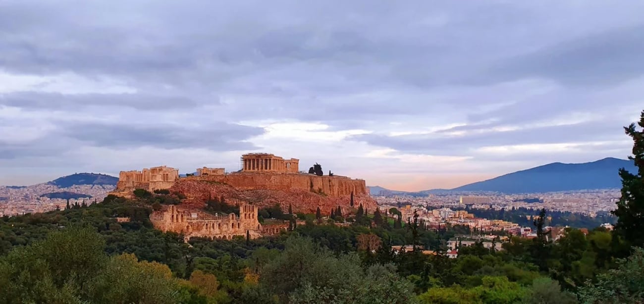 The Acropolis of Athens at sunset, with ancient ruins atop a rocky hill surrounded by greenery, city buildings in the background—reminding visitors that Athens is not just steeped in history but also stands among the biggest cities in the world—and a cloudy sky overhead.