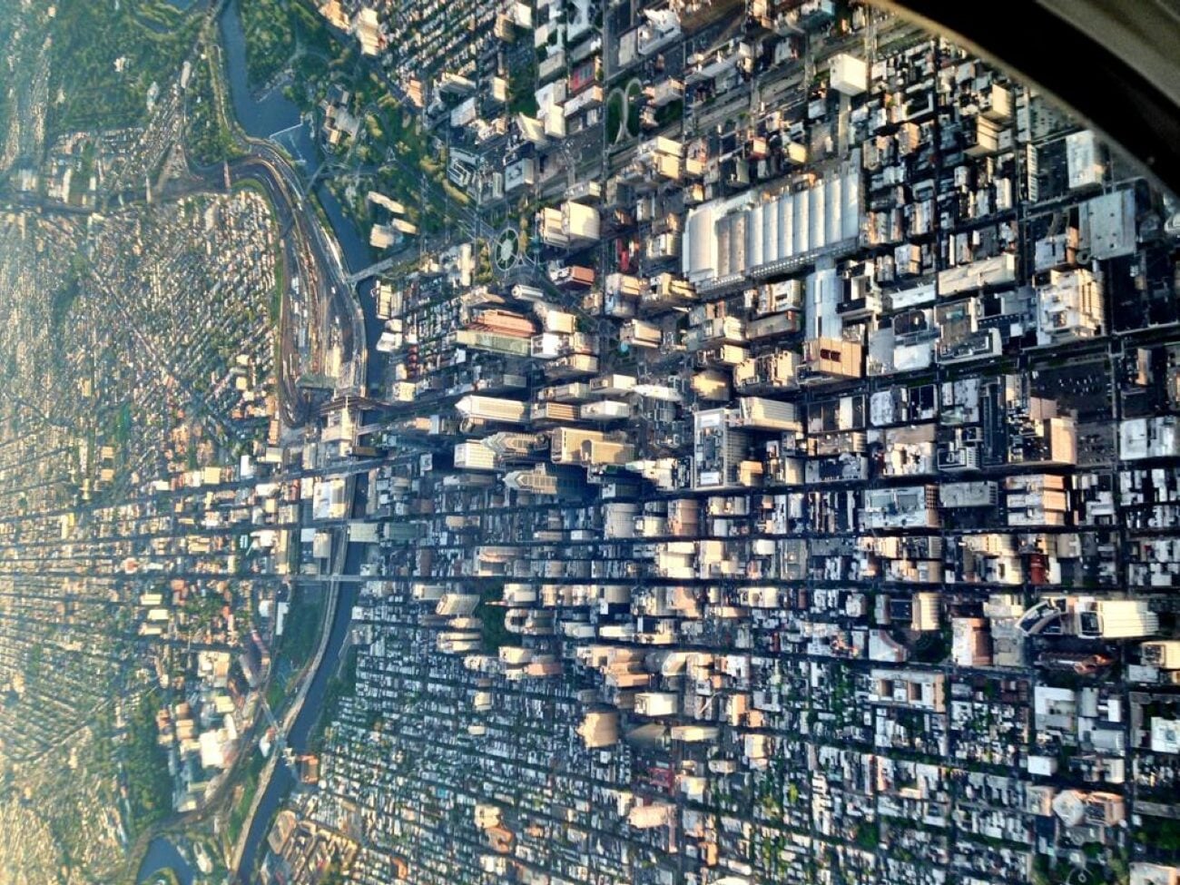 Aerial view of a dense urban cityscape with a grid of streets, tall buildings, and patches of greenery, as seen through airplane windows.